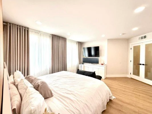 Modern bedroom with a large bed, white bedding, and multiple pillows. There are curtains covering windows, a flat-screen TV mounted on the wall, a white dresser, and a door with glass panels. The room has wooden flooring and recessed lighting.