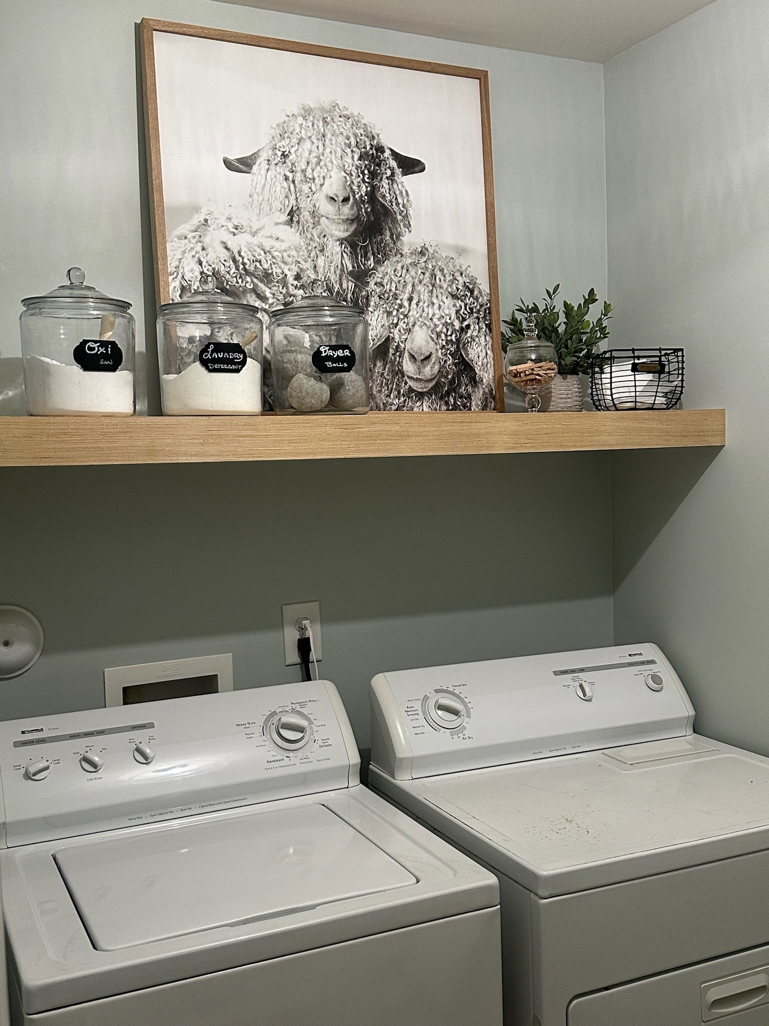 A laundry room with side-by-side washing machines under a wall-mounted wooden shelf. The shelf holds three labeled jars, a framed black-and-white photo of two sheep, a small potted plant, a jar with wooden clothespins, and a wire basket with folded i