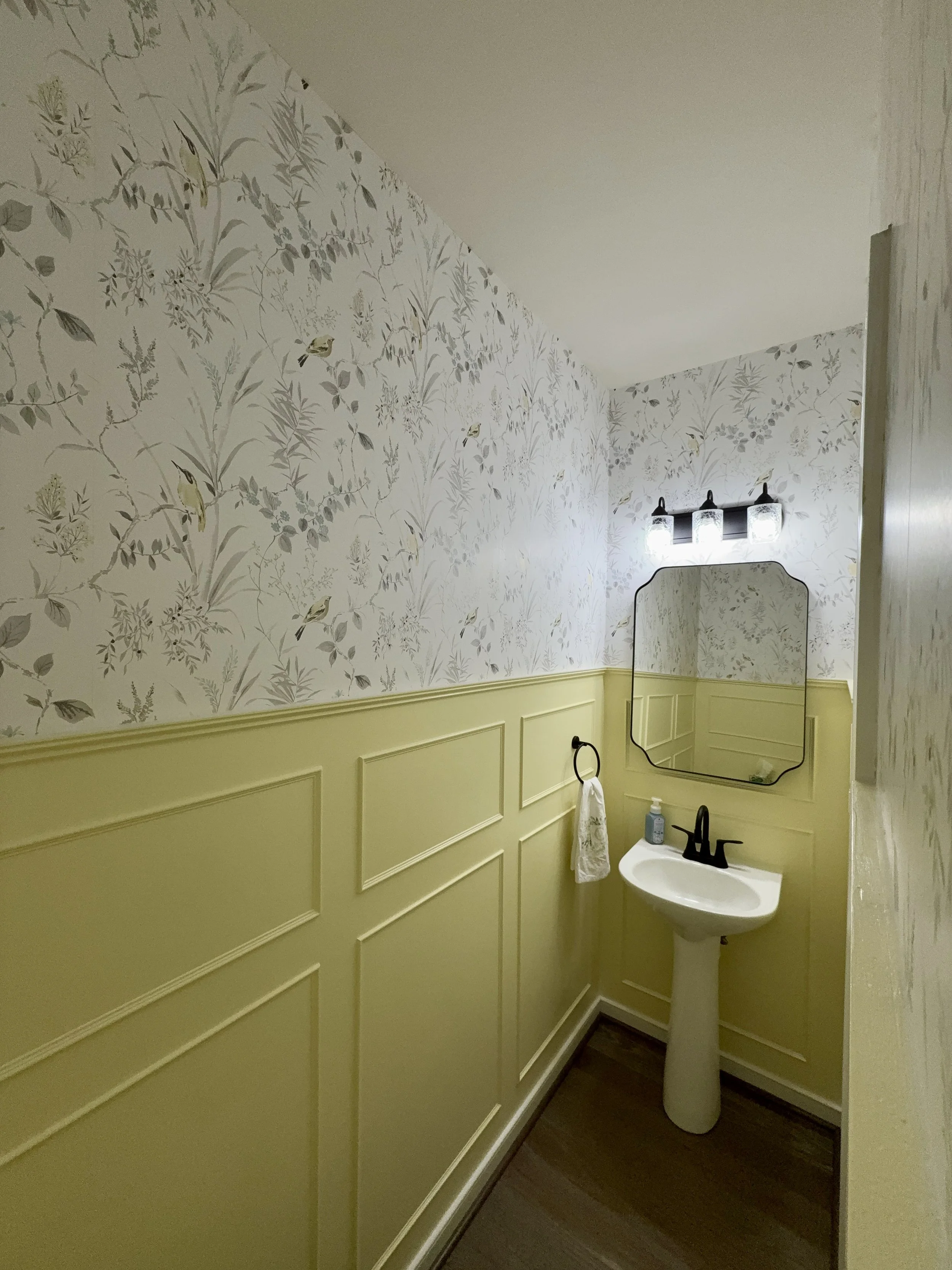 Small bathroom with a pedestal sink, black fixtures, and a rectangular mirror. The upper part of the walls is covered in floral wallpaper, while the lower part has yellow wainscoting. A black towel ring with a white hand towel and a soap dispenser ar