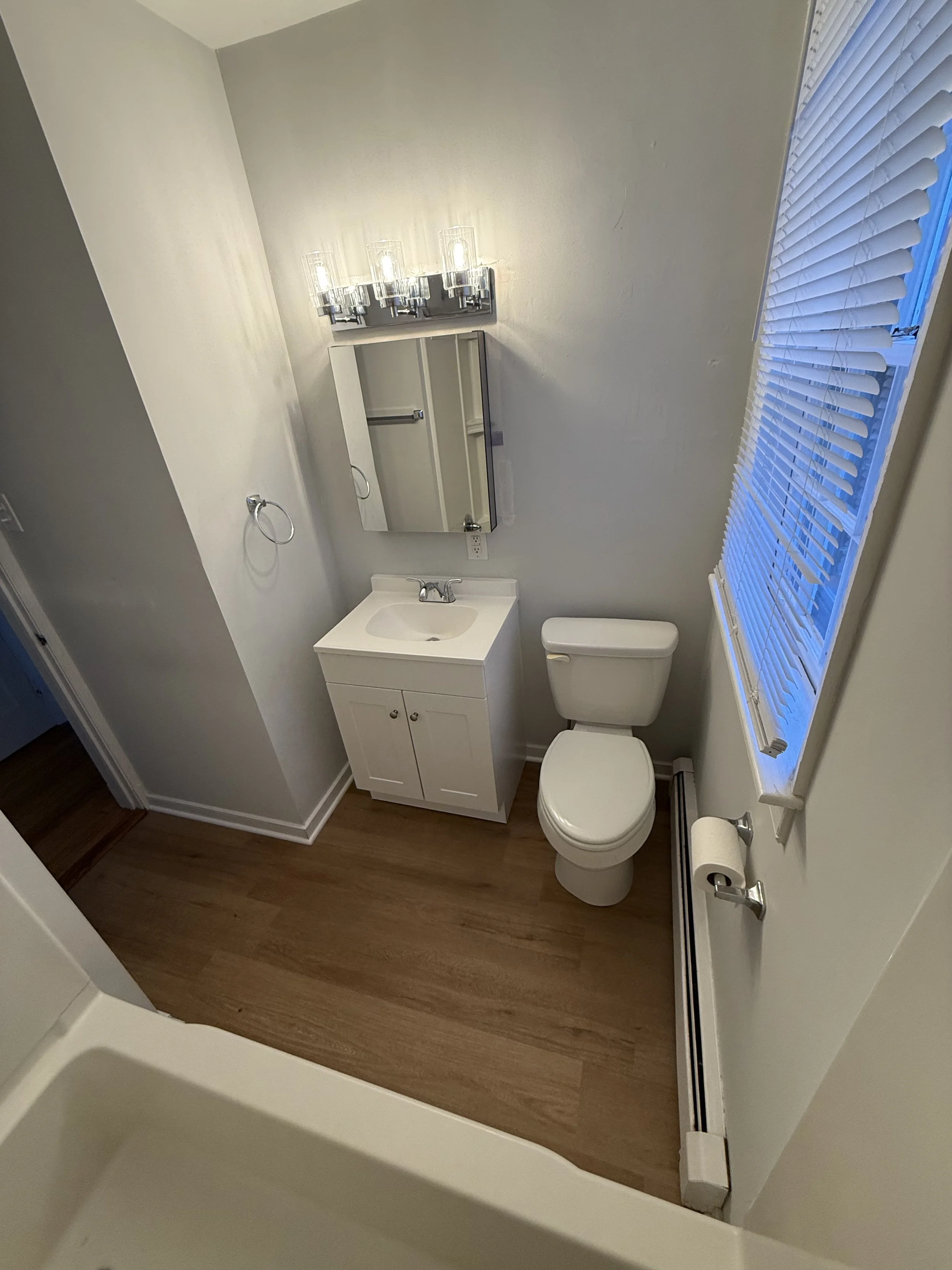 Small bathroom with a toilet, a small white vanity with a sink, a wall-mounted mirror, and a fixture with four clear glass lights above the mirror. There is a window with blinds, a towel ring on the wall, and a toilet paper holder on the wall next to