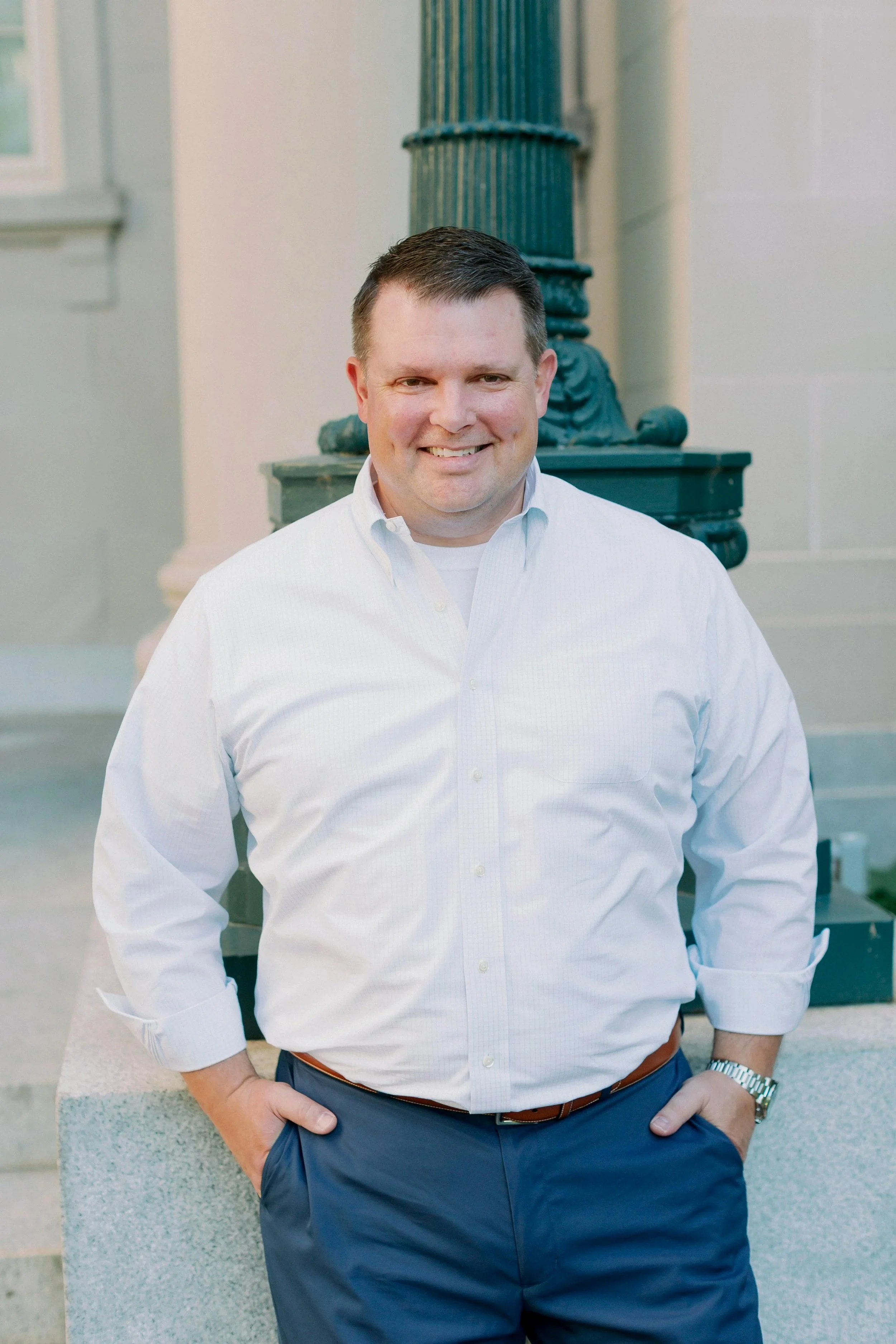 A man in a white button-up shirt and blue pants standing outdoors, smiling, with a decorative green post and building in the background.