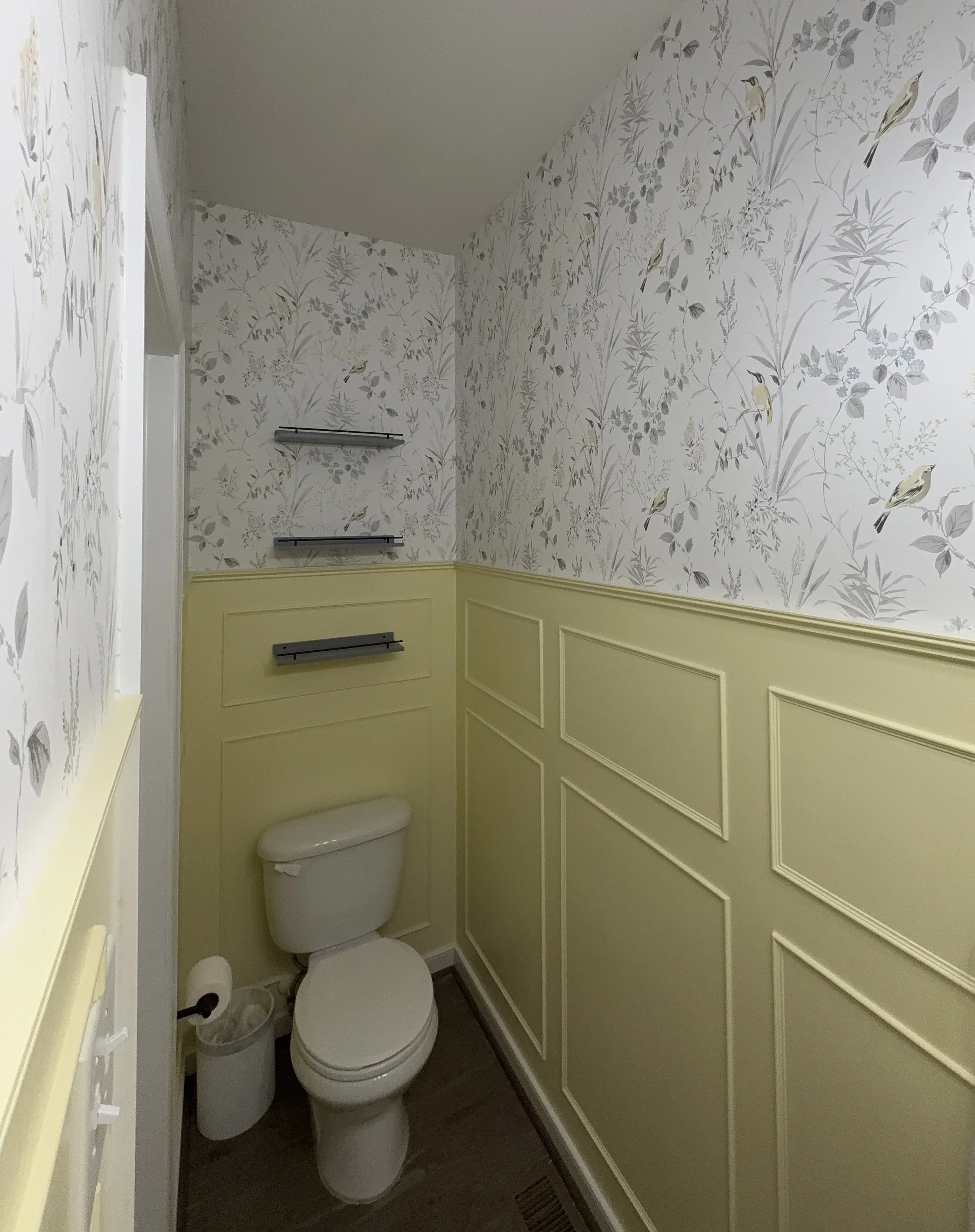 Small bathroom with yellow half-wall wainscoting, floral wallpaper, a toilet, and black shelves.