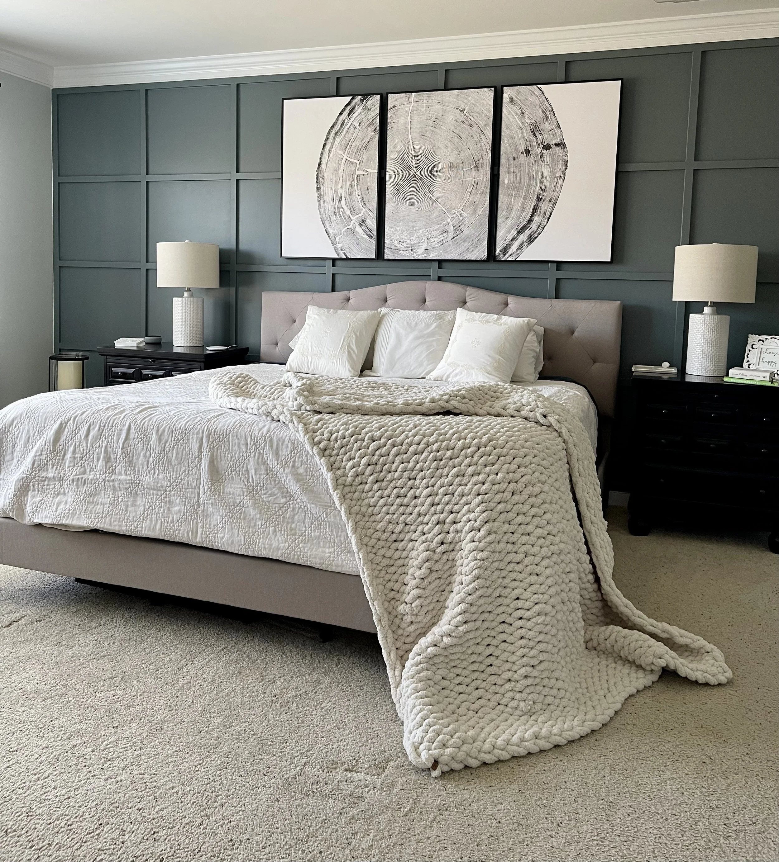 A cozy bedroom with a king-sized bed, white quilt, and plush chunky knit blanket. Two white pillows and a tufted beige headboard. Dark wood nightstands with white lamps on each side. A triptych art piece featuring a tree ring on the dark green panele