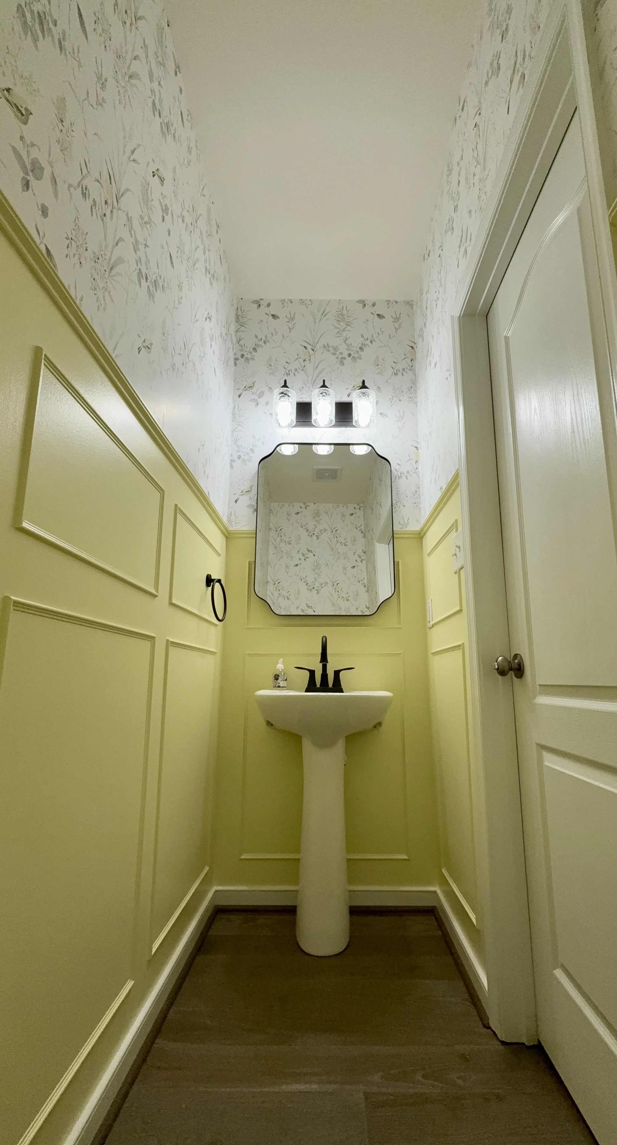 Small bathroom with pale yellow wainscoting, floral wallpaper, pedestal sink with black faucet, oval mirror, and three lights above the mirror.