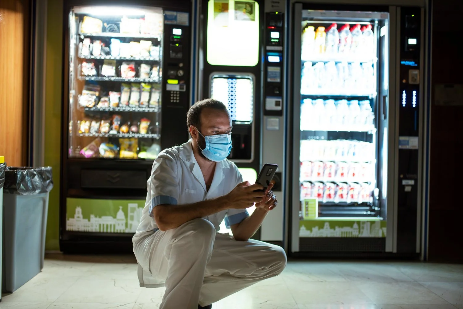 A man wearing a face mask and white clothes is crouching in front of vending machines, looking at his phone.