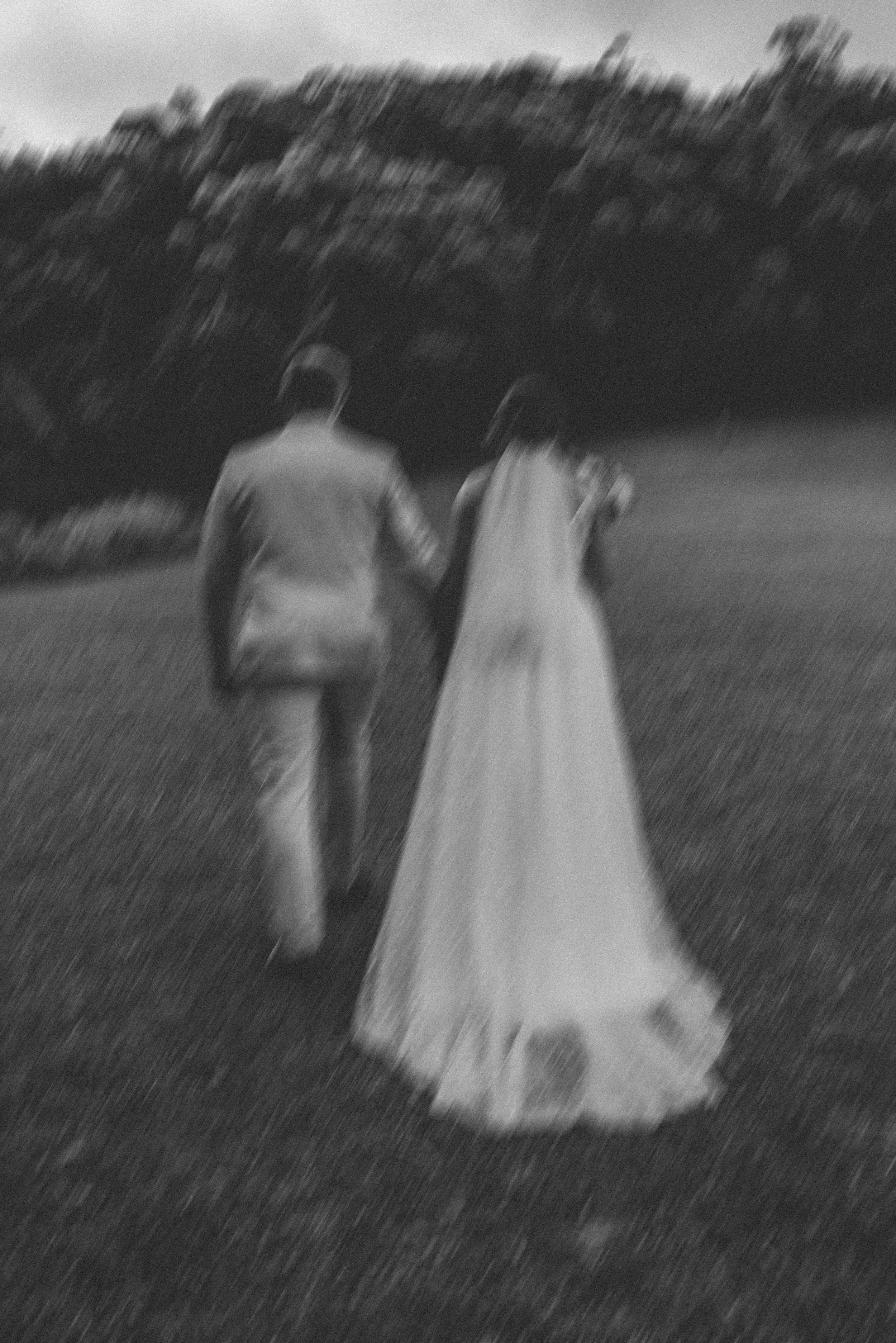 A black and white photo of a couple holding hands and walking together outdoors, with trees in the background.