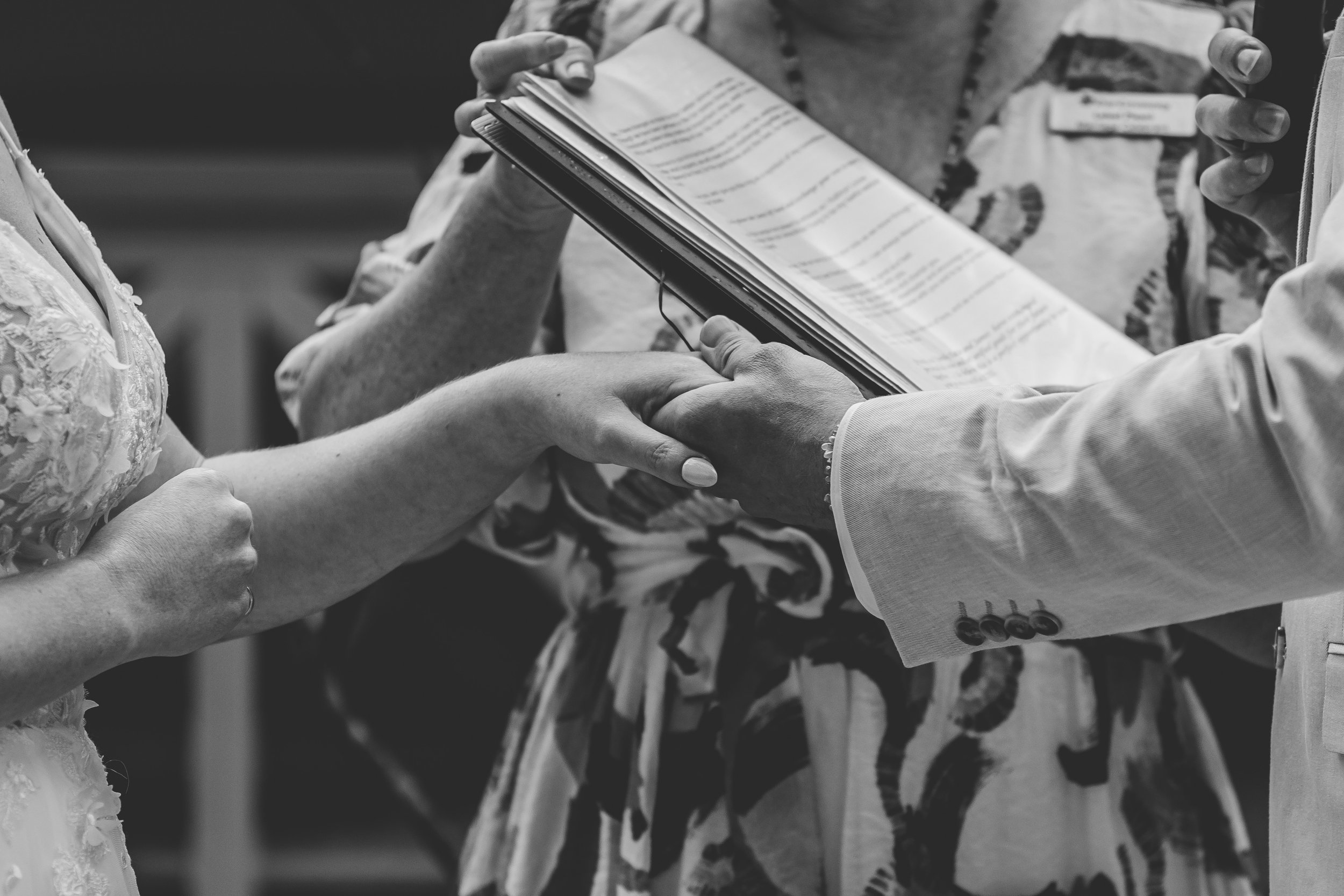 A wedding ceremony where a couple is exchanging vows, with a person officiating holding a book or script, and the bride's hand being held by the groom.