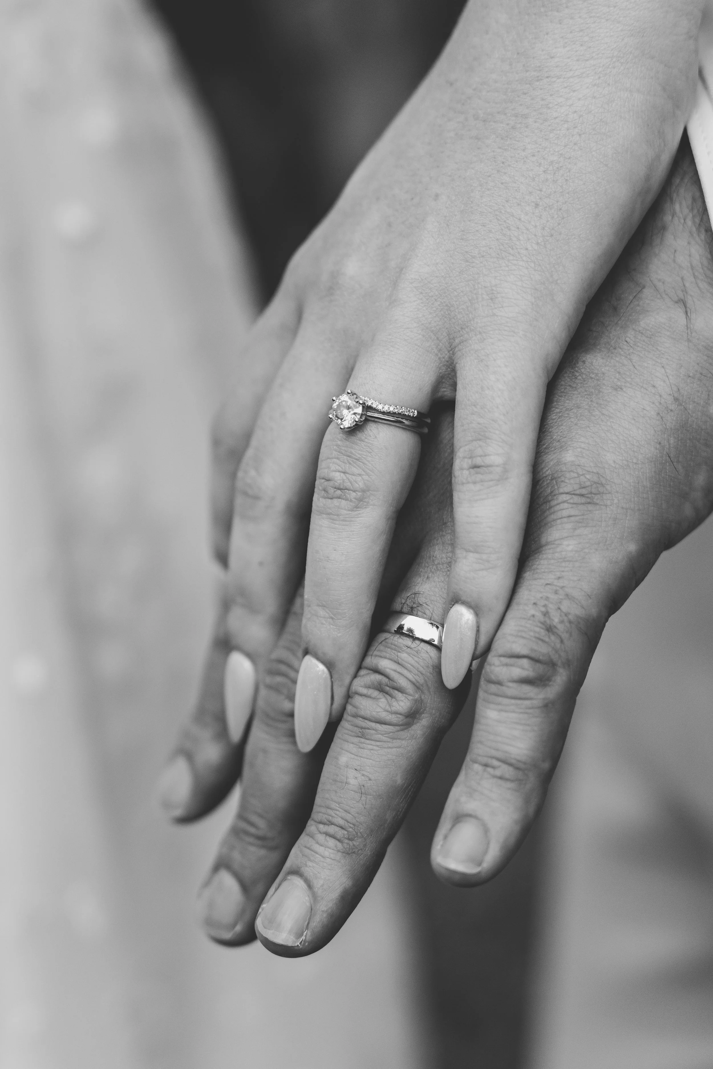 Close-up of a couple's hands showing their wedding rings, with the woman's hand on top of the man's hand, both with manicured nails and wedding bands in black and white.