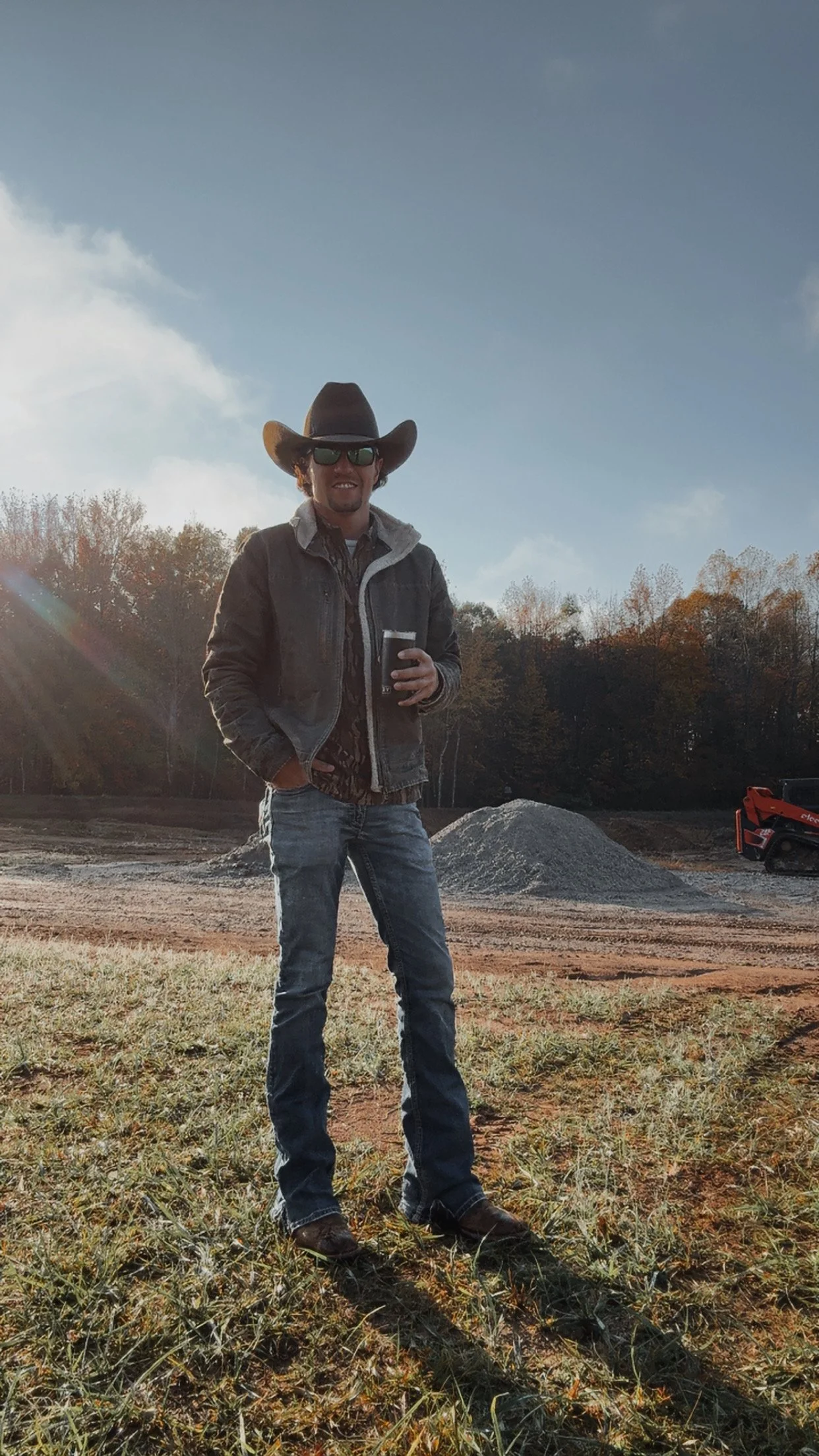 A man wearing a cowboy hat, sunglasses, and a denim jacket standing outdoors at a construction or excavation site during late afternoon, holding a drink.