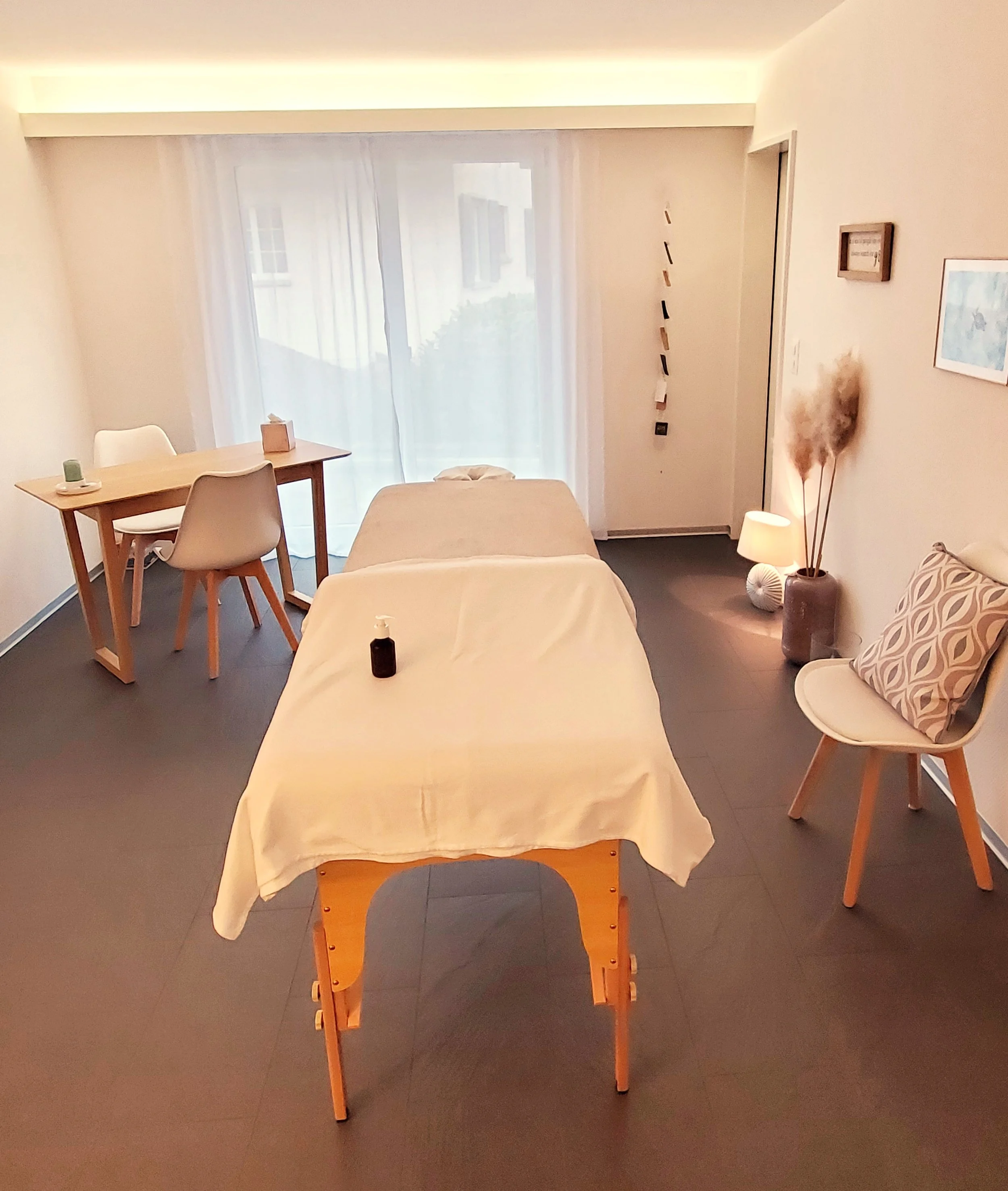 Ein hell erleuchteter Raum mit einem Behandlungsbett in der Mitte, auf dem eine Lotion steht. An der Rückwand befindet sich ein Tisch mit zwei Stühlen, eine kleine Lampe steht auf einem Gartenzwerg, und es gibt einen Balkon mit Vorhängen. An der Wand hängen Bilder und eine dekorative Kette.