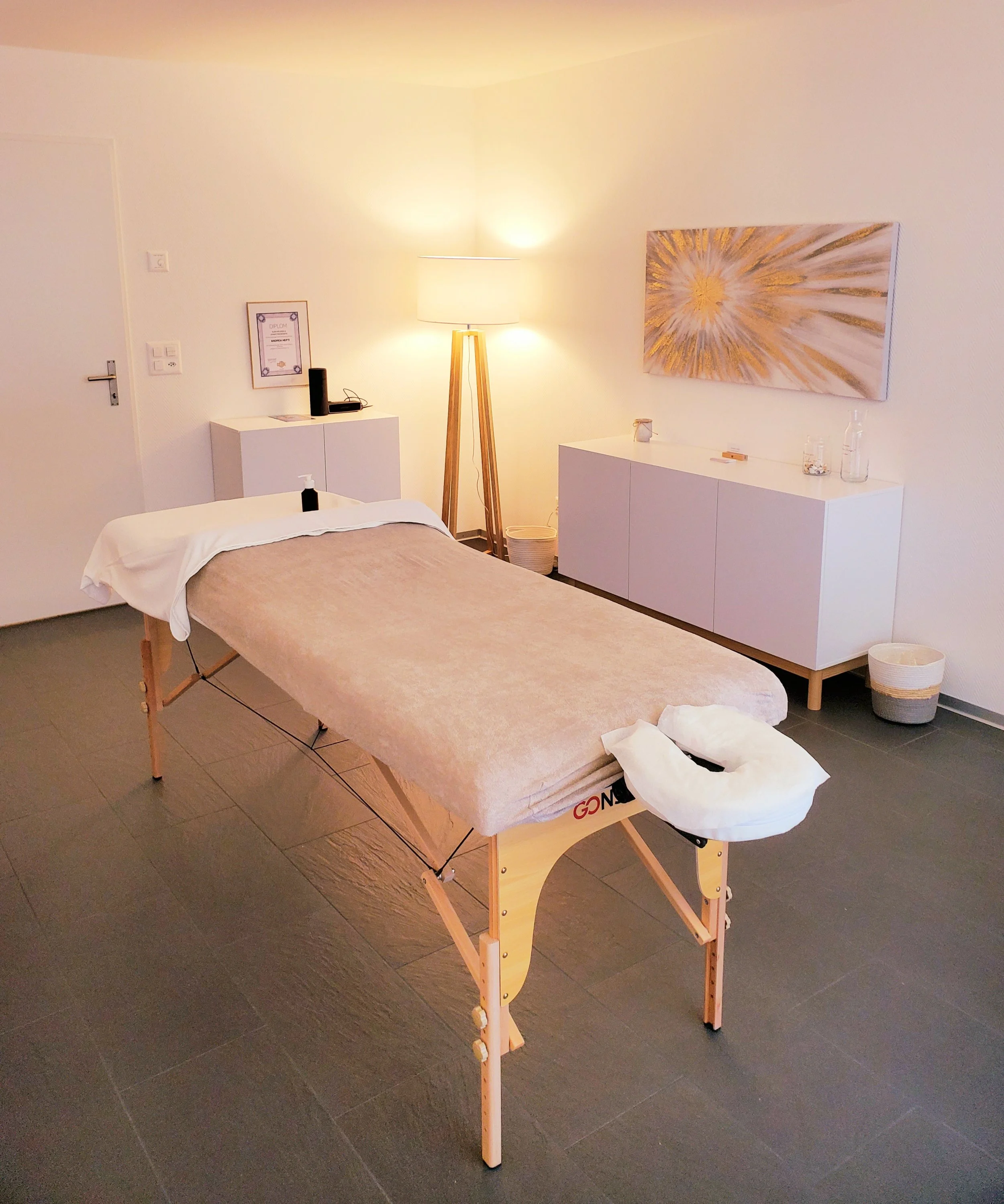 Behandlungszimmer mit Massagetisch, seitlicher Deckenlampe, weißen Möbeln und moderner Kunst an der Wand.