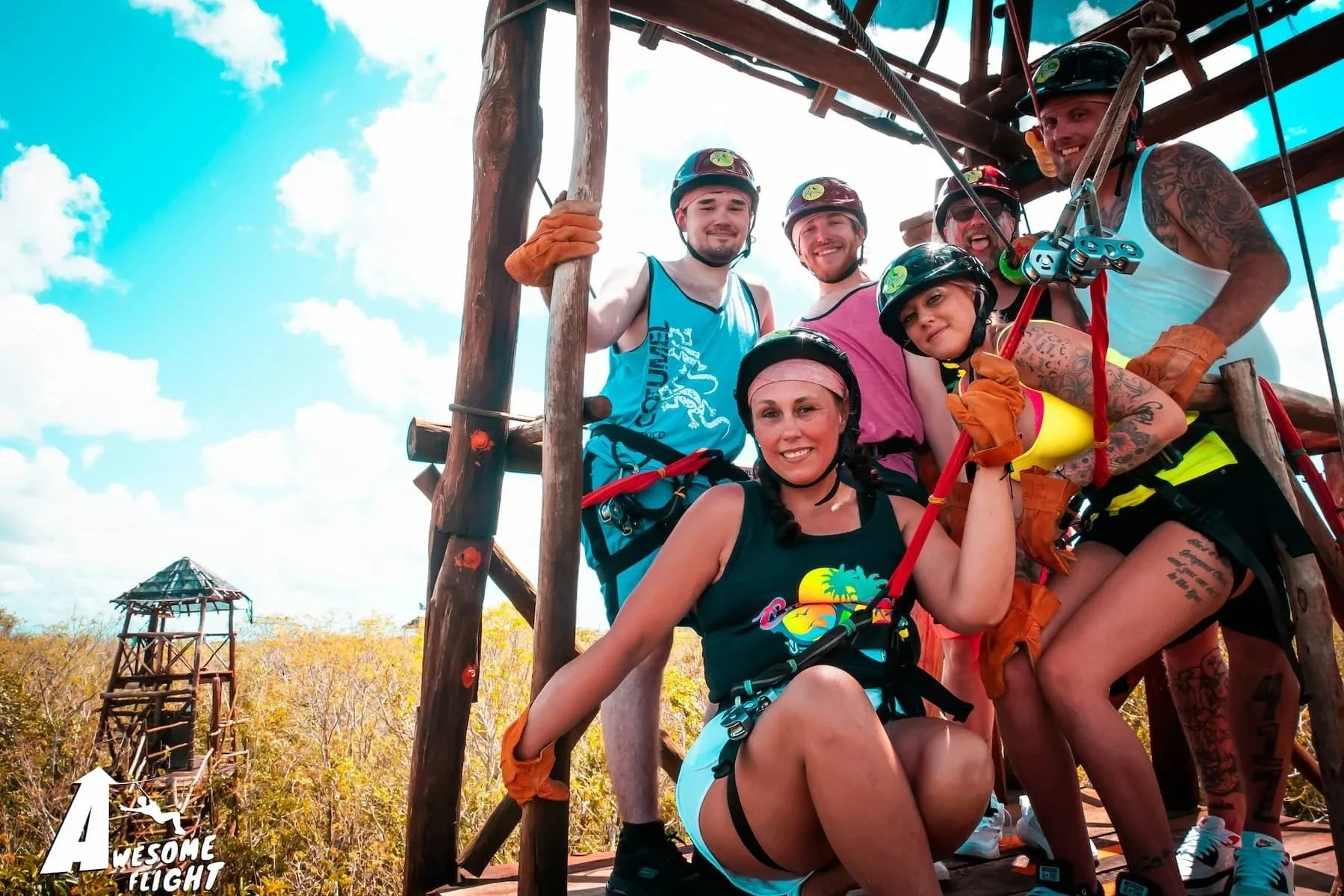Group of six people wearing helmets and harnesses, smiling and posing on a wooden platform for zip-lining, with a lush forest and a zip line tower in the background on a sunny day.