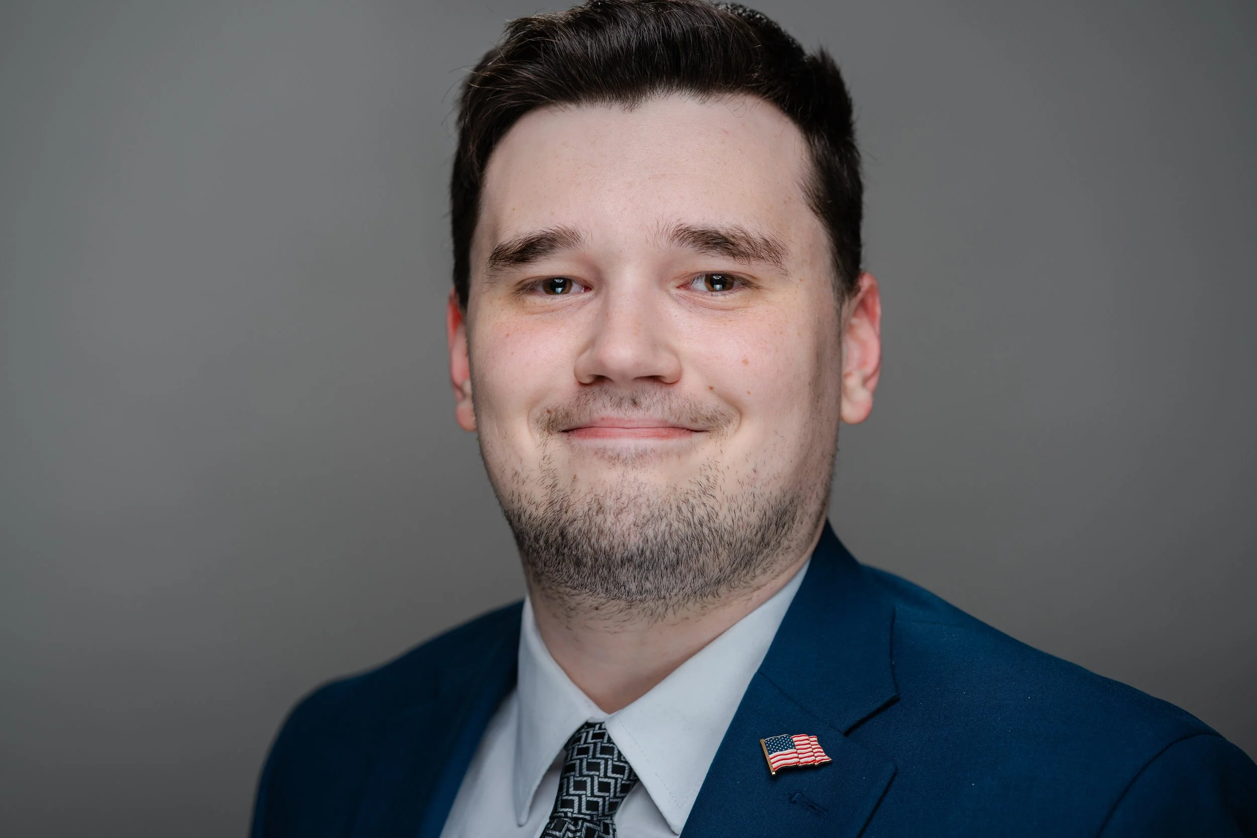 Close-up of a smiling young man with dark hair, wearing a blue suit, white shirt, and a patterned tie, with a small American flag lapel pin, against a gray background.