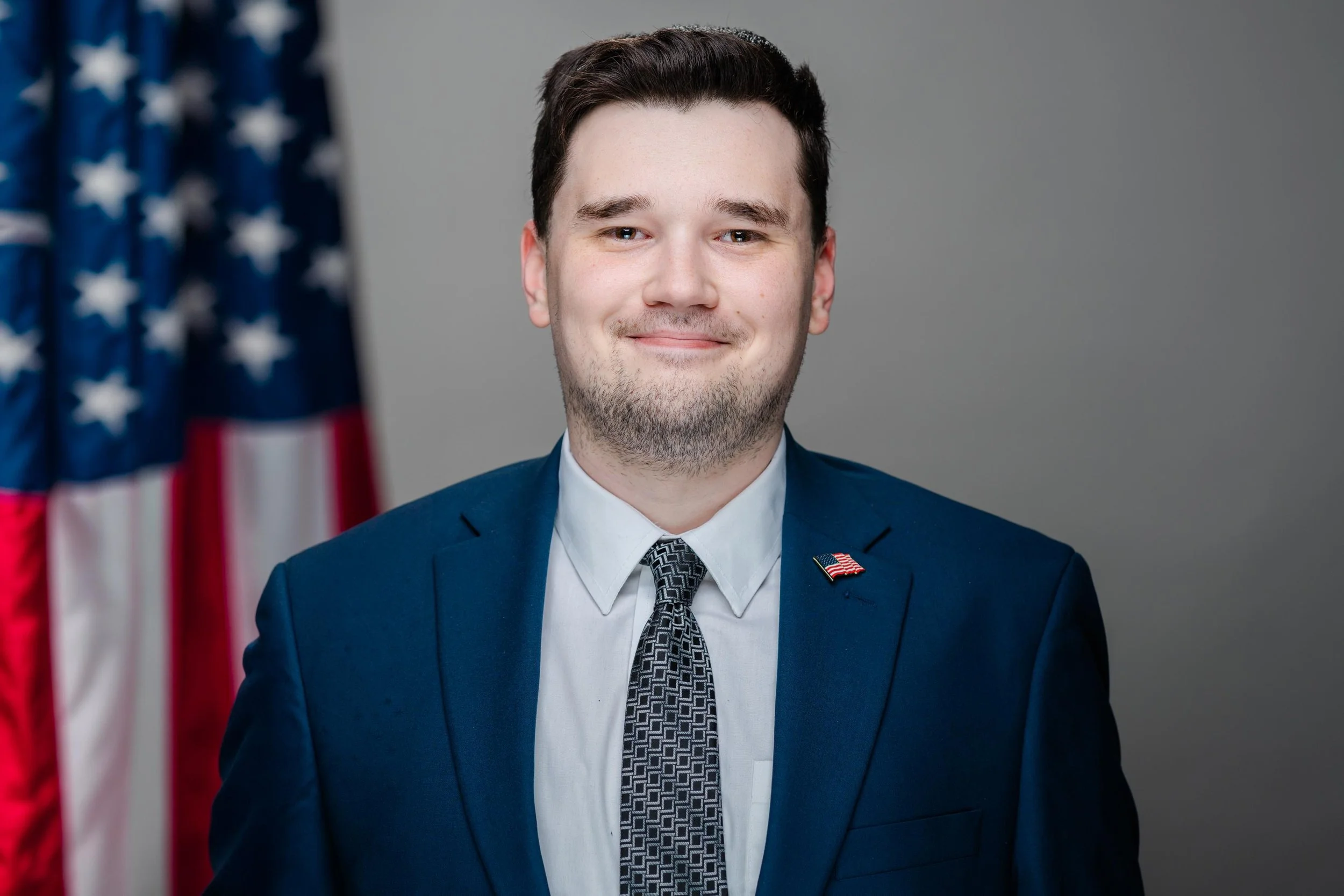 Portrait of a young man in a navy blue suit and flag lapel pin, smiling with an American flag in the background.