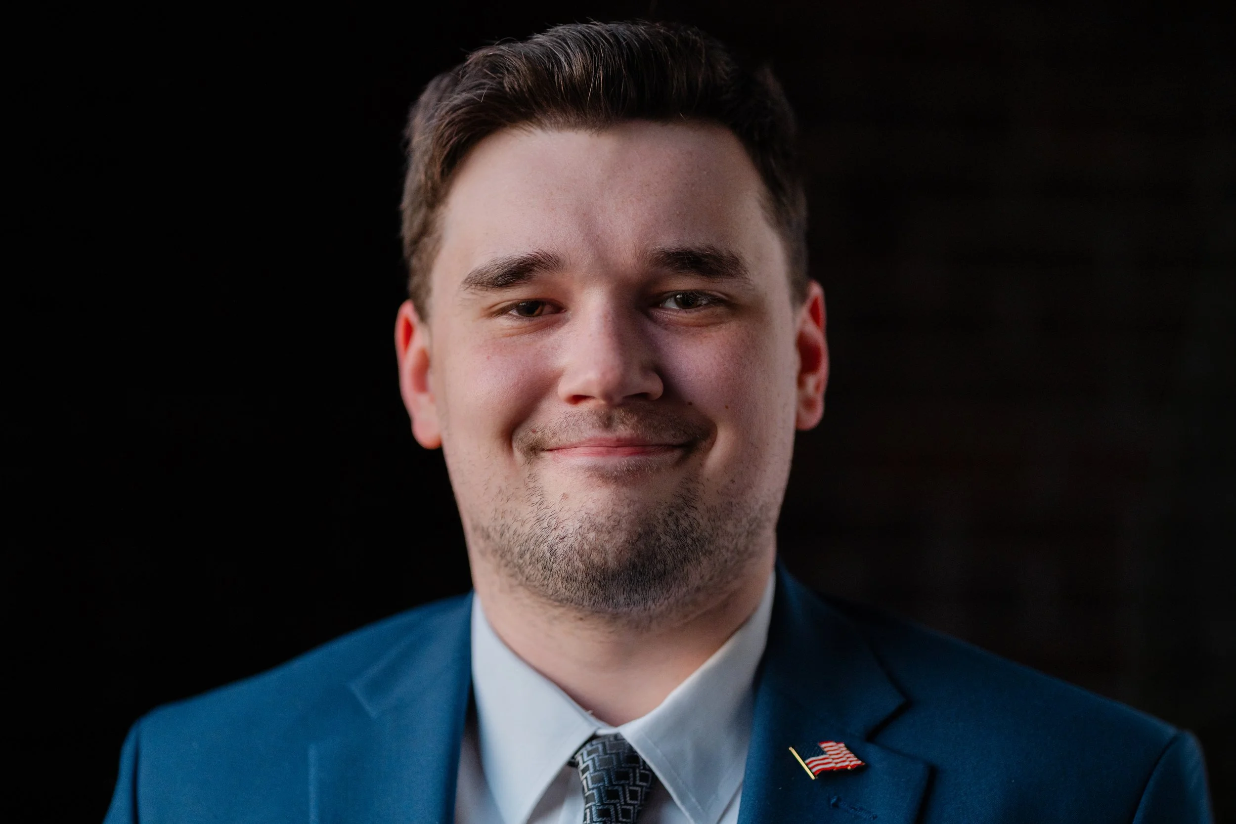Close-up of a young man with short dark hair, wearing a blue suit, white shirt, black patterned tie, and lapel pin with American flag, smiling against a dark background.