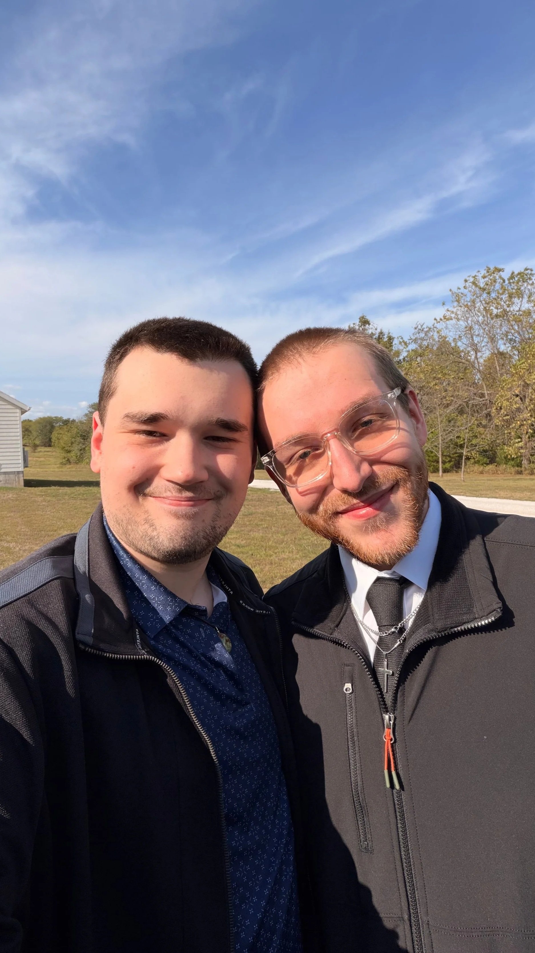 Two smiling men take a selfie outdoors on a sunny day, with one wearing glasses, a cross necklace, a black jacket, and the other wearing a black jacket, with trees and a blue sky in the background.