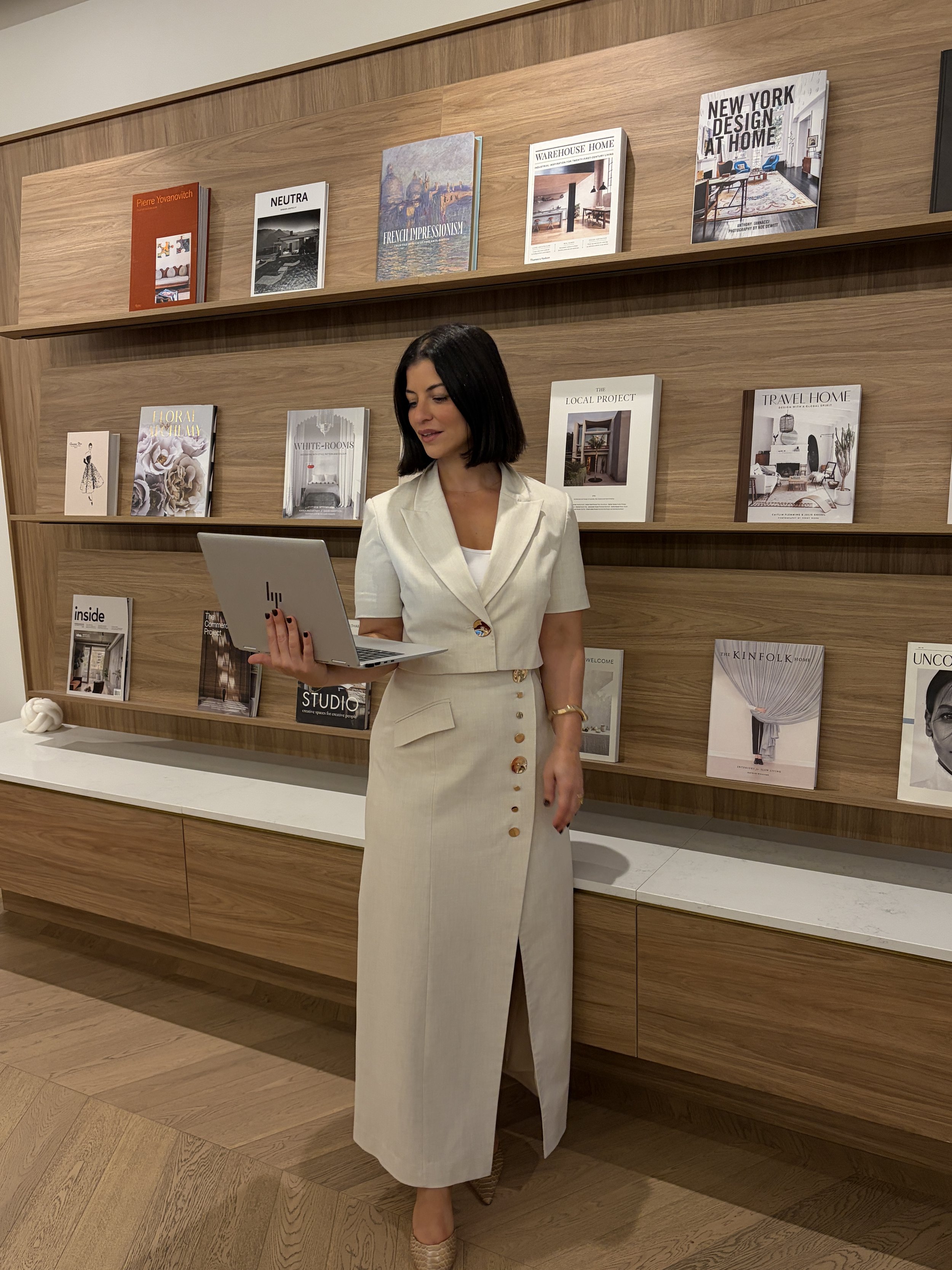 A woman with dark hair wearing a cream-colored suit with buttons, holding a laptop, standing in front of a wooden bookshelf with various magazines.