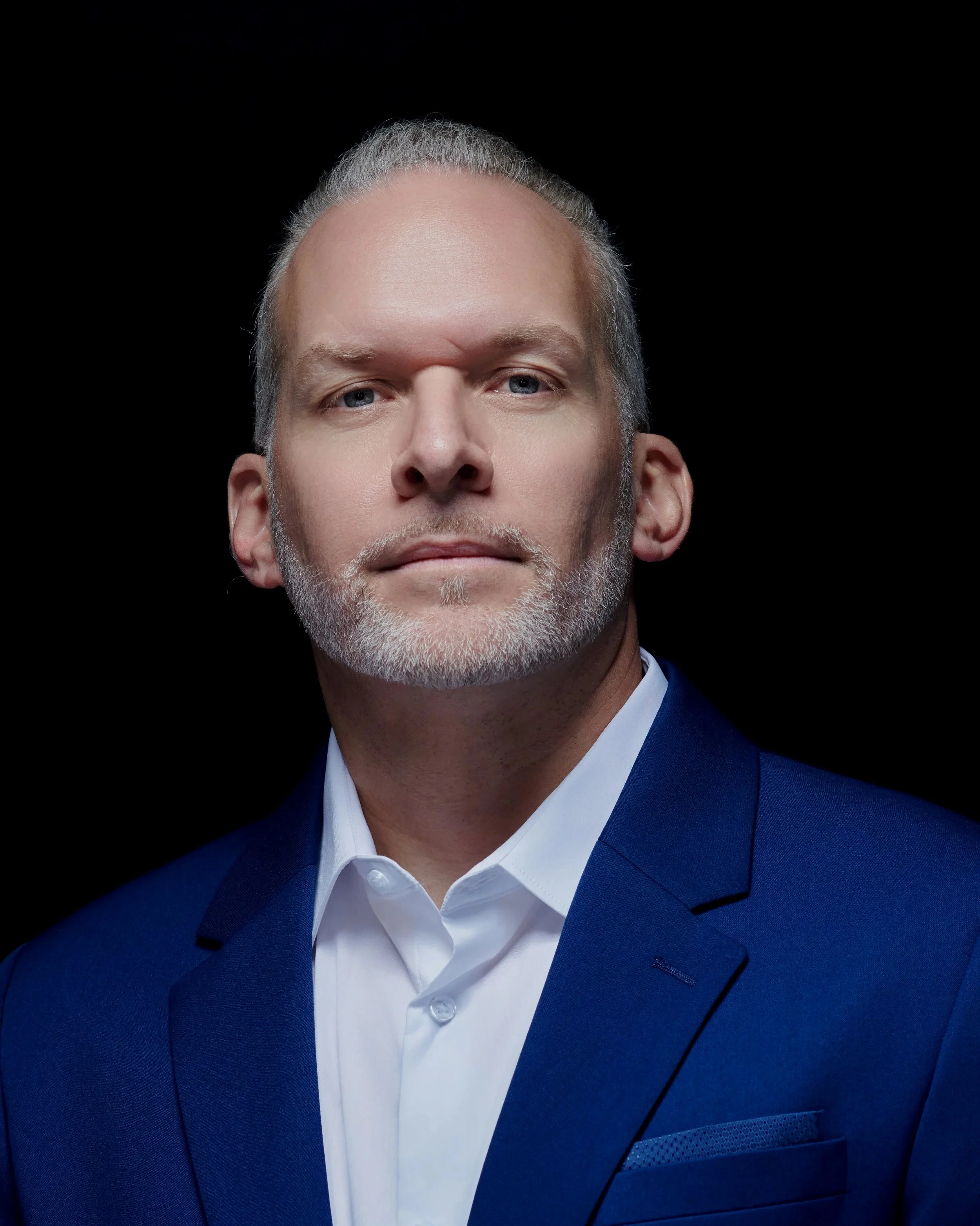 Homme d'âge moyen avec une barbe blanche, portant un costume bleu et une chemise blanche, regardant droit devant contre un fond noir.