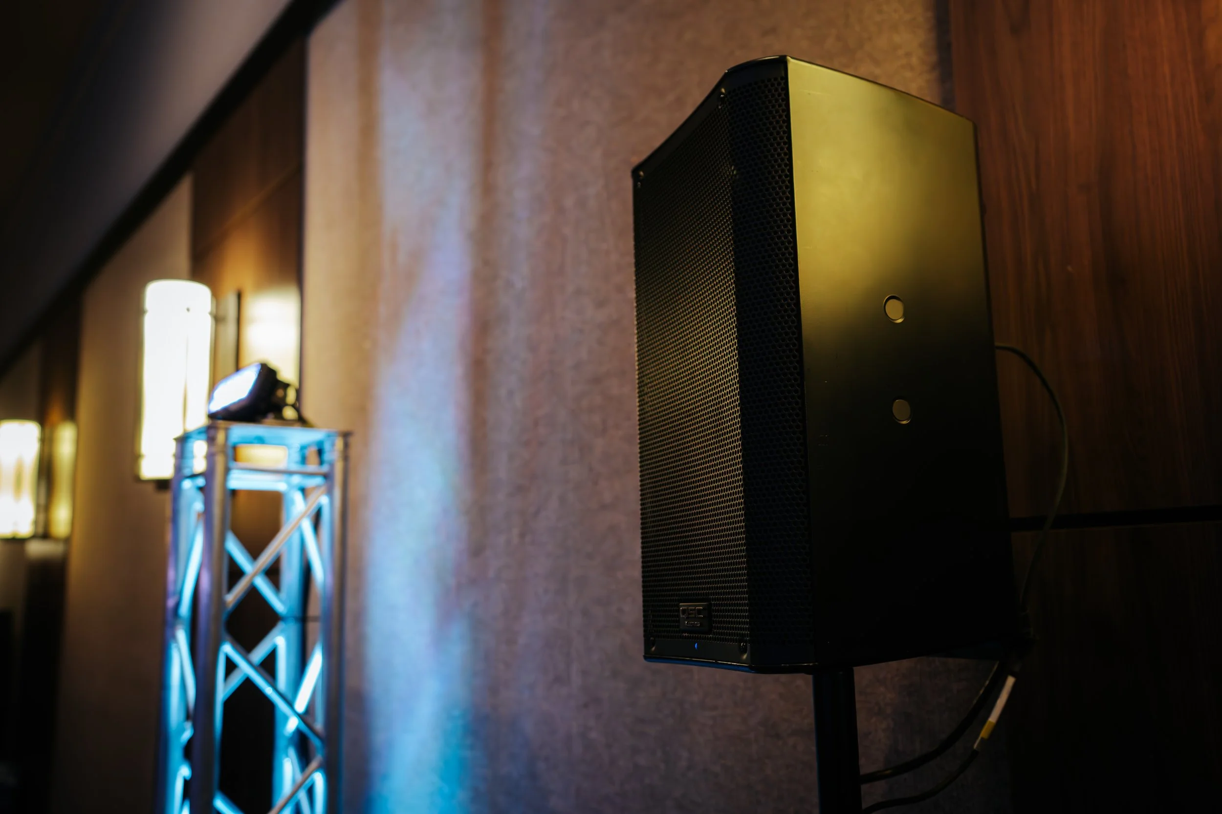 Enceinte noire montée sur un support, avec un mur en bois derrière, éclairée par des lumières de scène dans un espace intérieur.