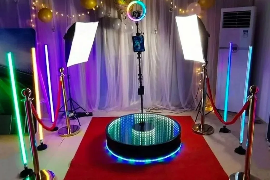 Zone photo avec un miroir rond, éclairage LED coloré, deux lumières d'éclairage, tapis rouge, barrière en corde rouge, cercle lumineux au sol, et décoration lumineuse en arrière-plan.