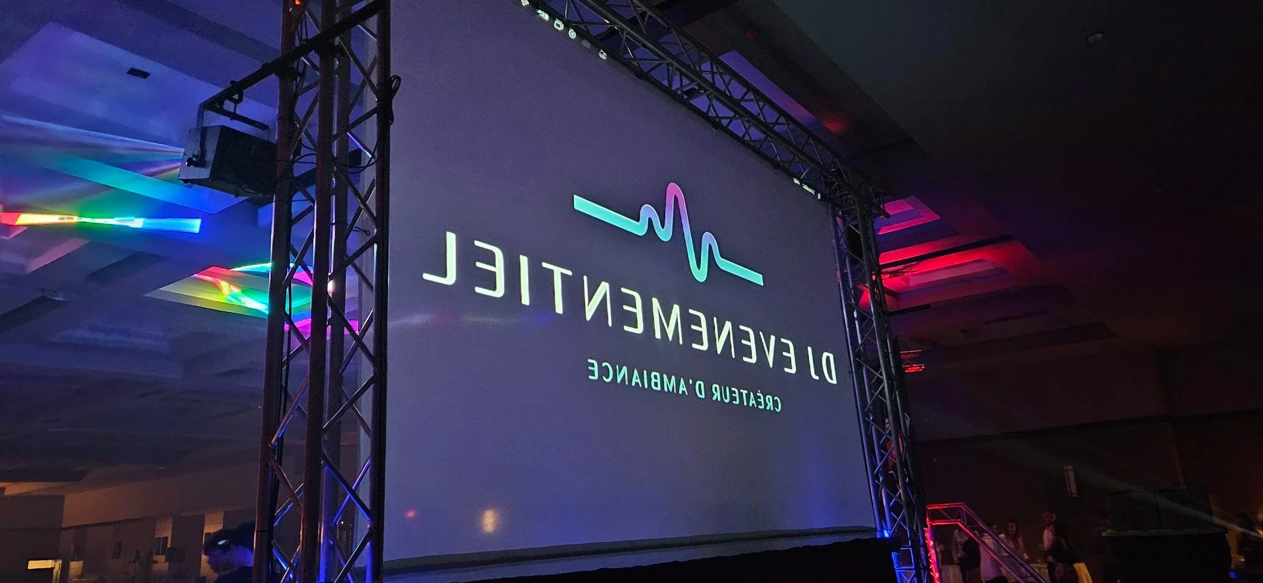 Grand écran affichant un logo avec une ligne de battements de cœur et le texte 'DEVENTEIL CRETEUR D'AMBIANCE' à l'intérieur d'une salle de conférence, avec des lumières colorées et des personnes en arrière-plan.