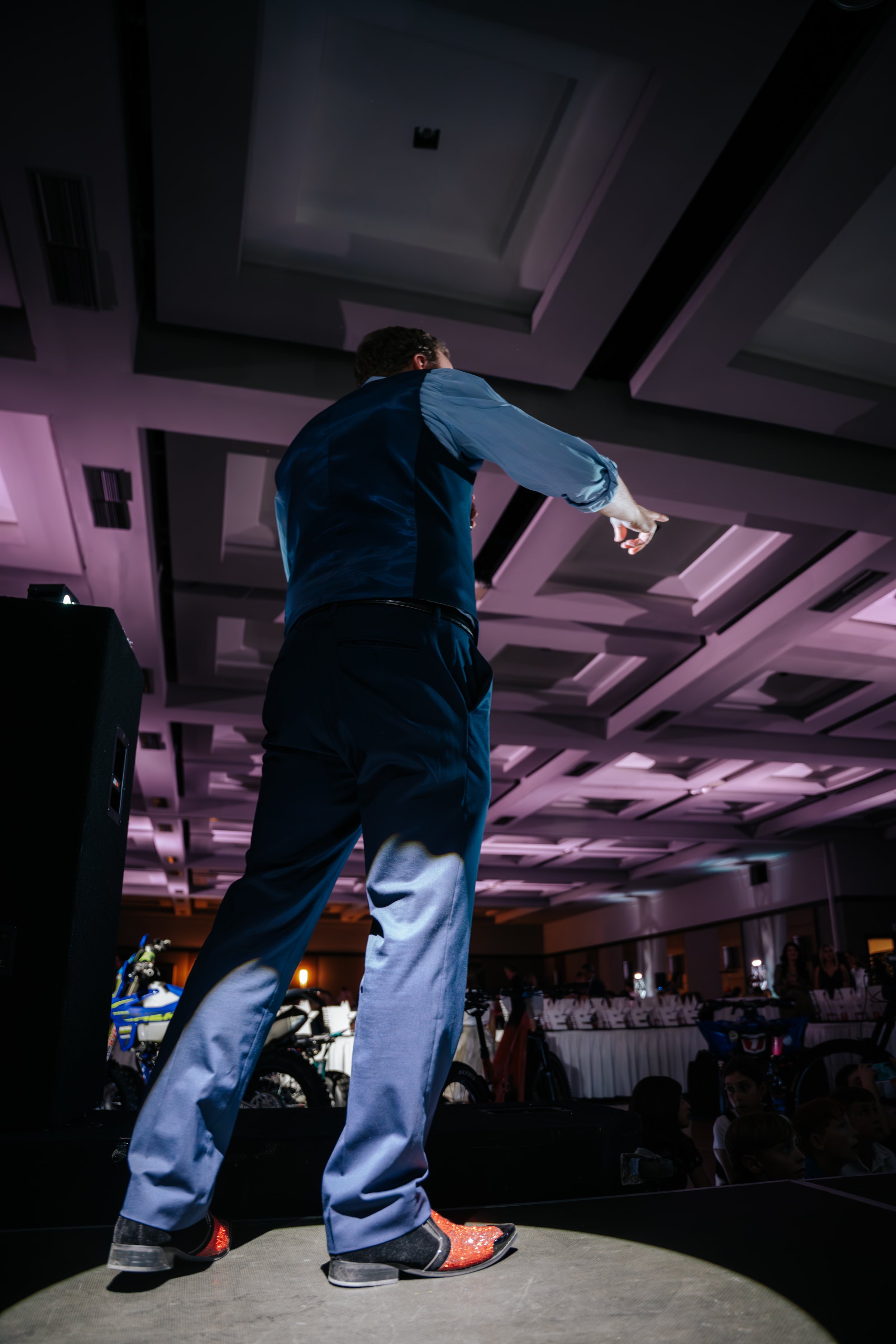 Un homme en costume sombre, vu de dos, qui pointe du doigt vers la droite, se tenant sur scène dans une salle de réception ou un grand hall, avec des enfants et d'autres personnes au fond.