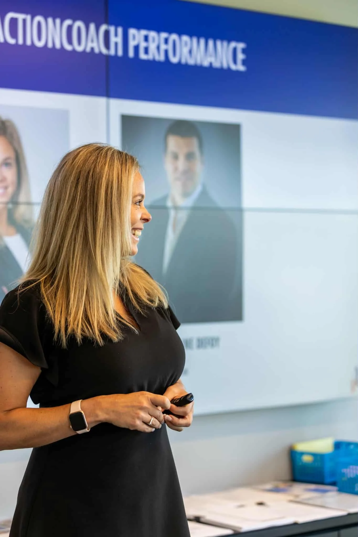 Femme souriante regardant à gauche, portant une montre connectée, dans une salle de conférence avec un écran affichant 'ACTIONCOACH PERFORMANCE'