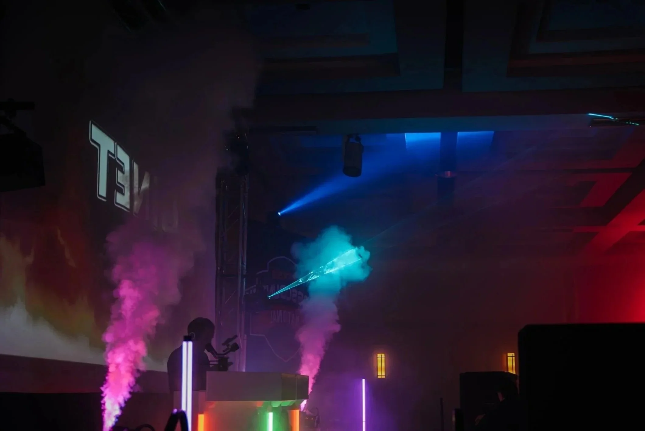 une scène de concert ou spectacle avec des effets de fumée colorée, des projecteurs bleus et roses, un DJ ou un artiste en arrière-plan, décor lumineux avec des colonnes de lumières vertes, et un logo 'T3V' projeté en haut à gauche.