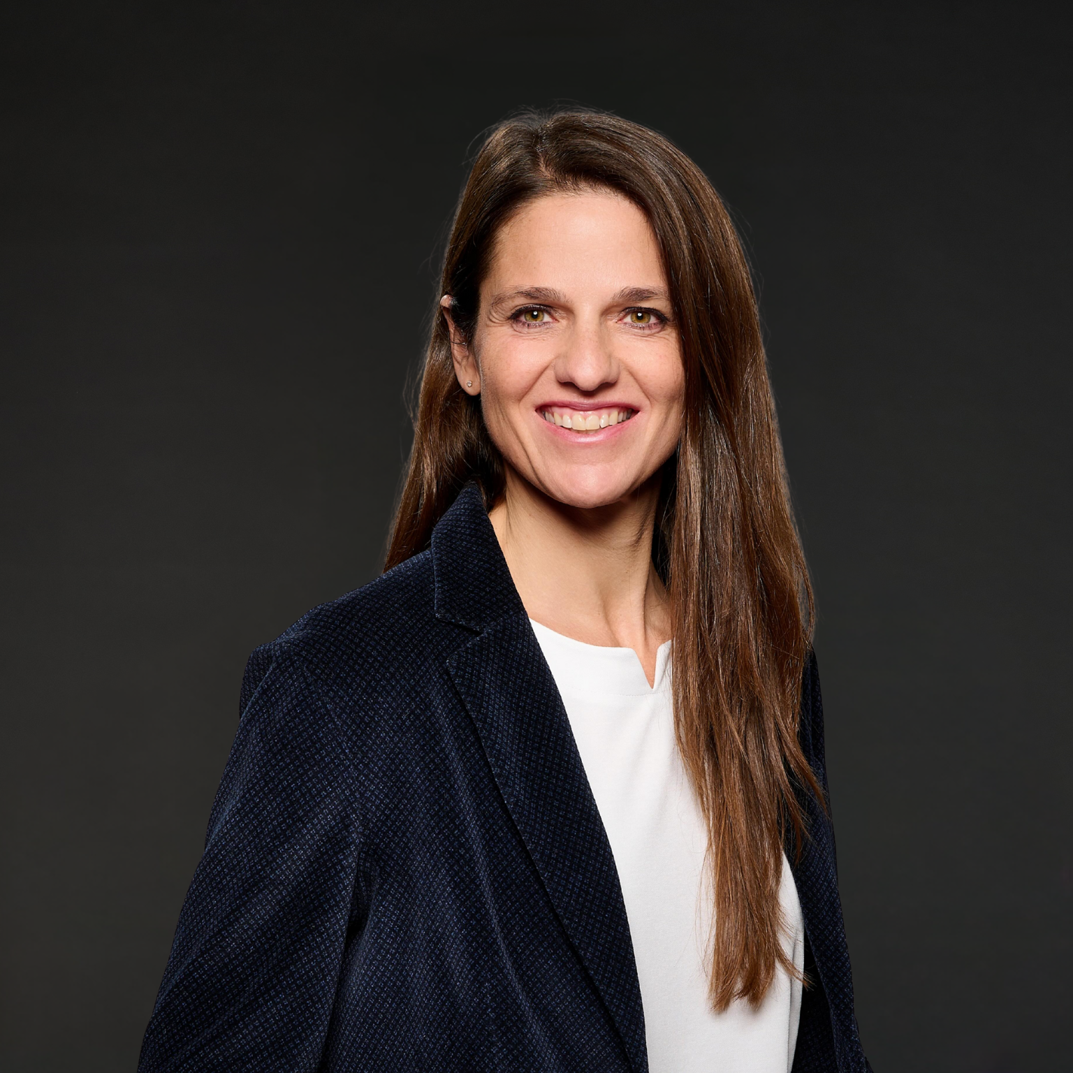 Eine Frau mit langem braunem Haar, lächelnd, trägt eine dunkelblaue Jacke und ein weißes Oberteil, vor dunklem Hintergrund.
