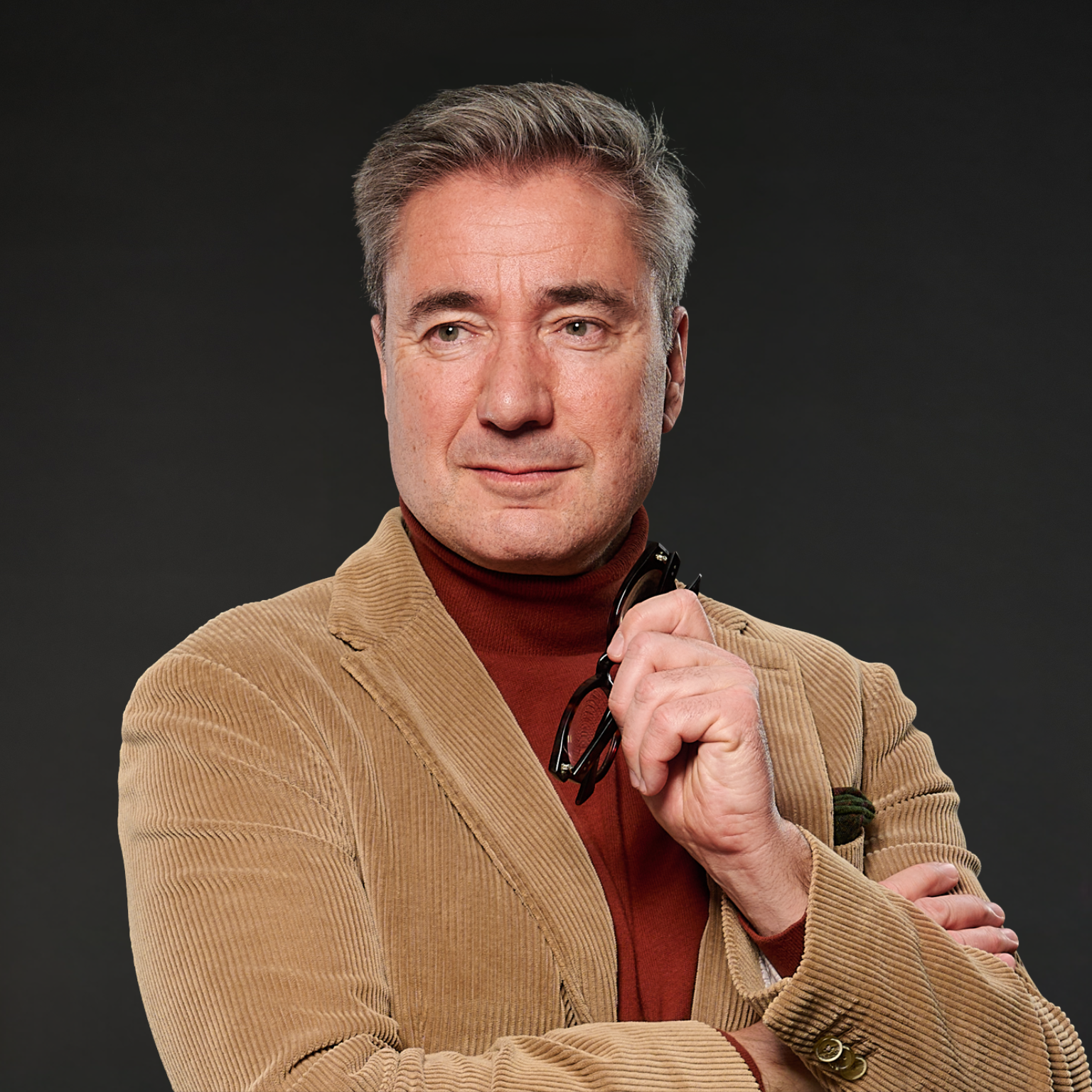 Ein Mann mit kurzen grauen Haaren, trägt einen braunen Cordjacke und einen roten Pullover. Er hält eine Brille in der rechten Hand und blickt nach links. Der Hintergrund ist dunkel.