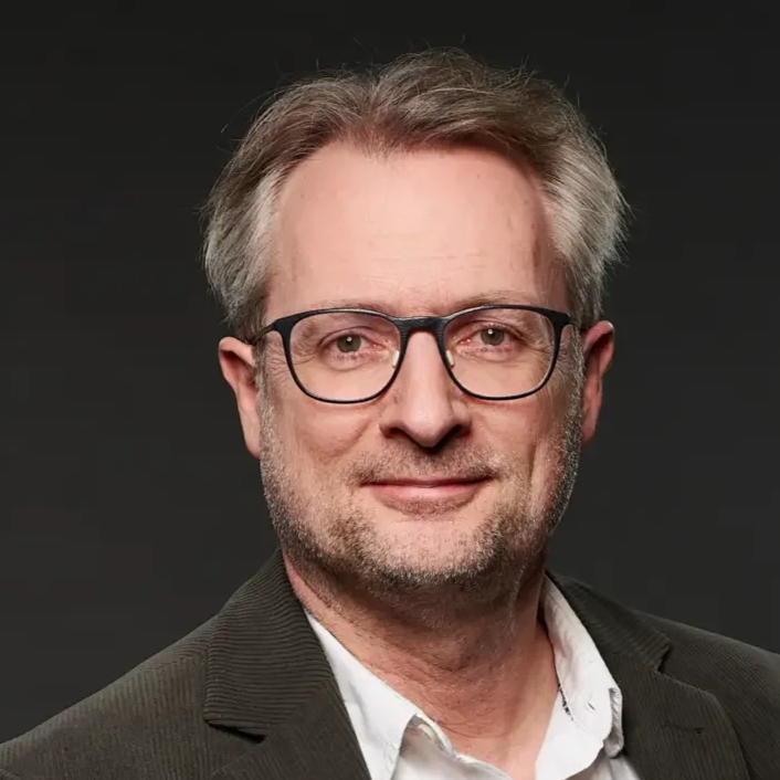 Porträt eines Mannes mit Brille, graues Haar und grauem Bart, in einem dunklen Hintergrund.