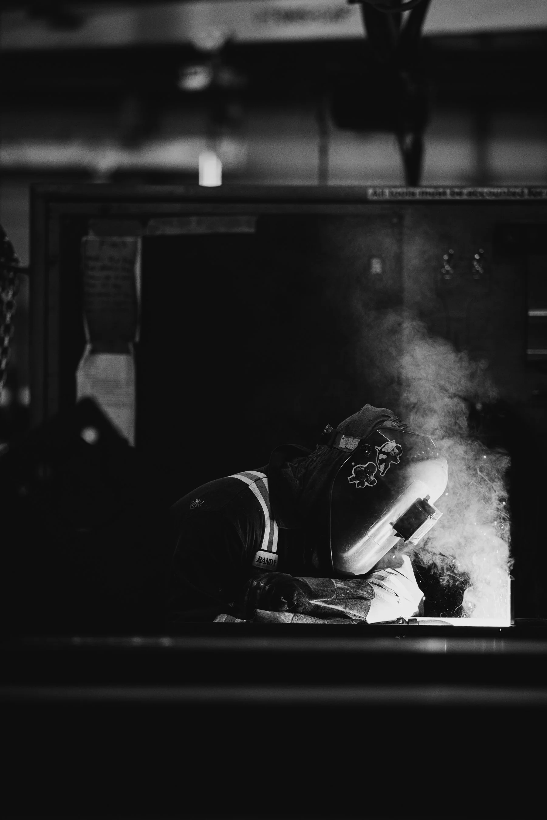 A welder wearing protective gear, including a welding mask with cartoon character decals, is working with bright sparks and smoke in a dark workshop.