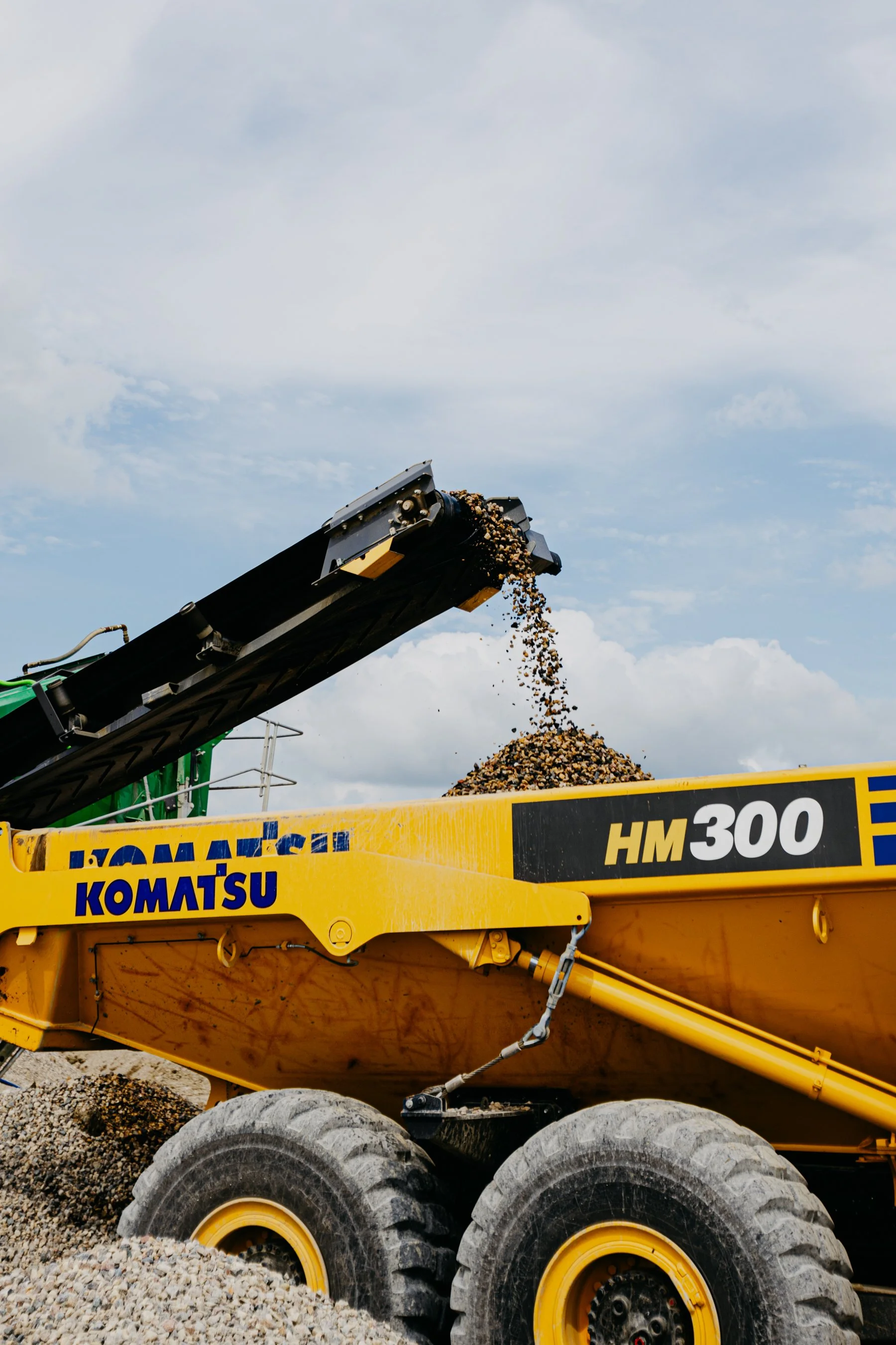 A yellow Komatsu HM300 heavy-duty dump truck unloading a load of gravel or rocks at a construction or mining site, with cloudy sky in the background.
