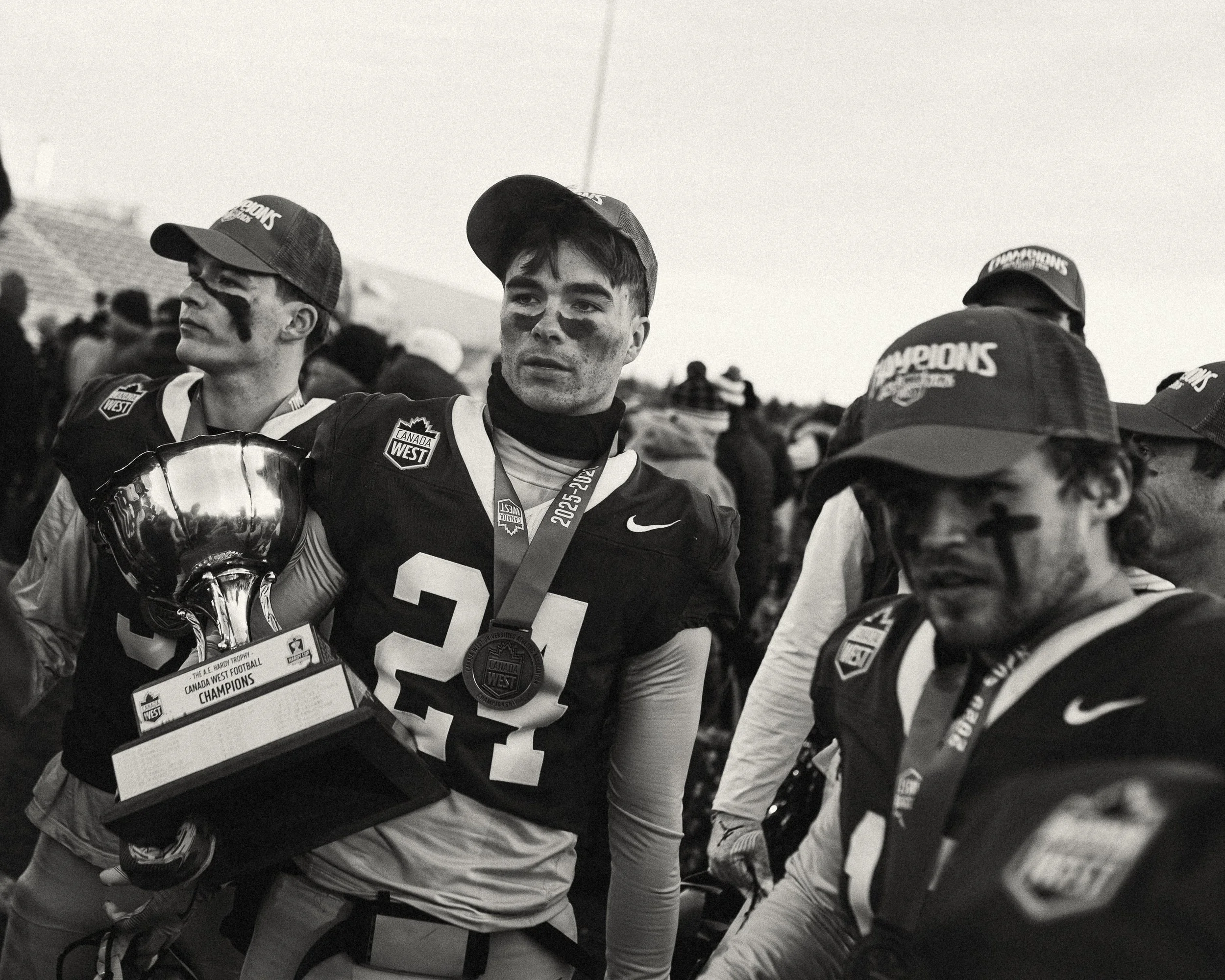 Black and white photo of football players celebrating with a trophy and medals, wearing caps and jerseys with 'Champions' and 'Canada' logos, in a crowd setting.