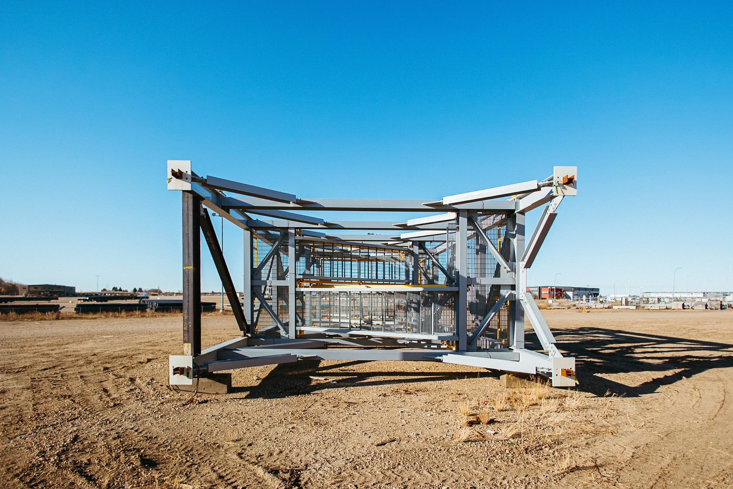 A large metallic industrial structure with a rectangular frame on an arid, sandy lot under a clear blue sky.