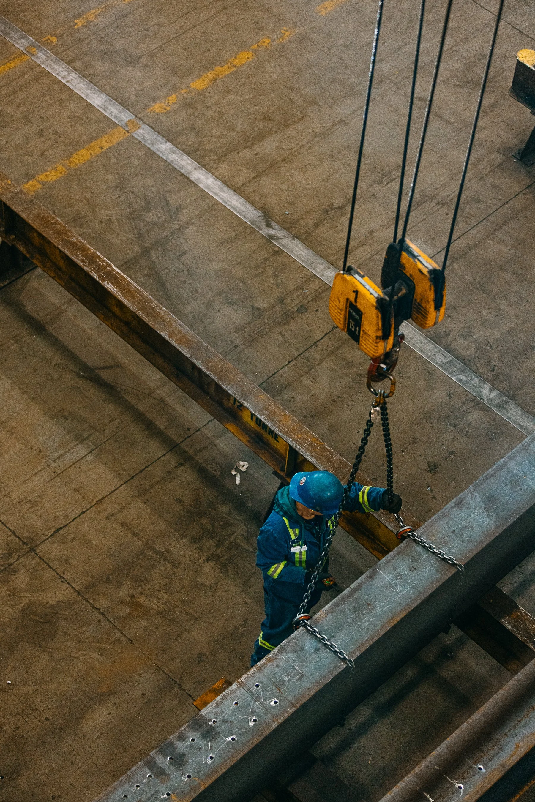 A worker wearing a blue safety helmet and safety gear is working inside a construction site with metal beams. The worker is suspended by chains attached to a hoist, and the site features a concrete floor with some yellow painted lines.