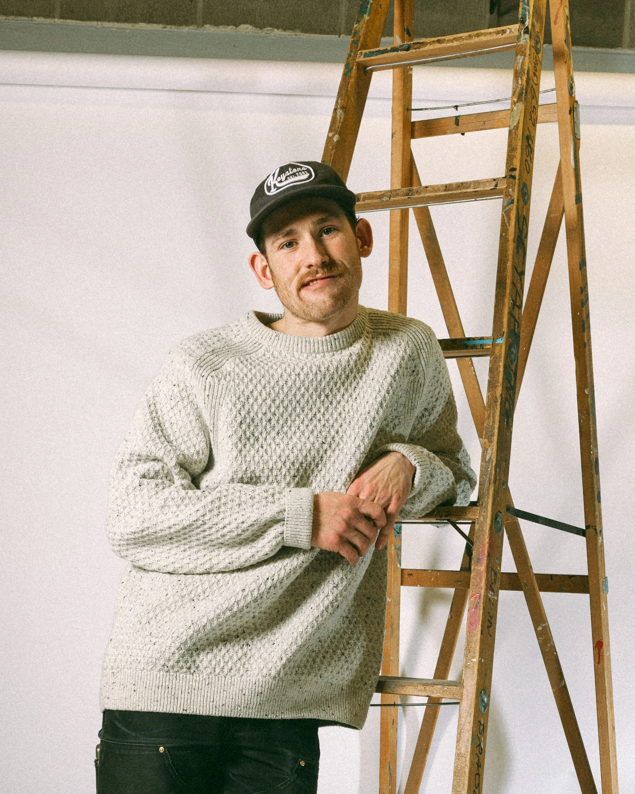 A man in a cream-colored sweater and black cap standing next to a wooden ladder.