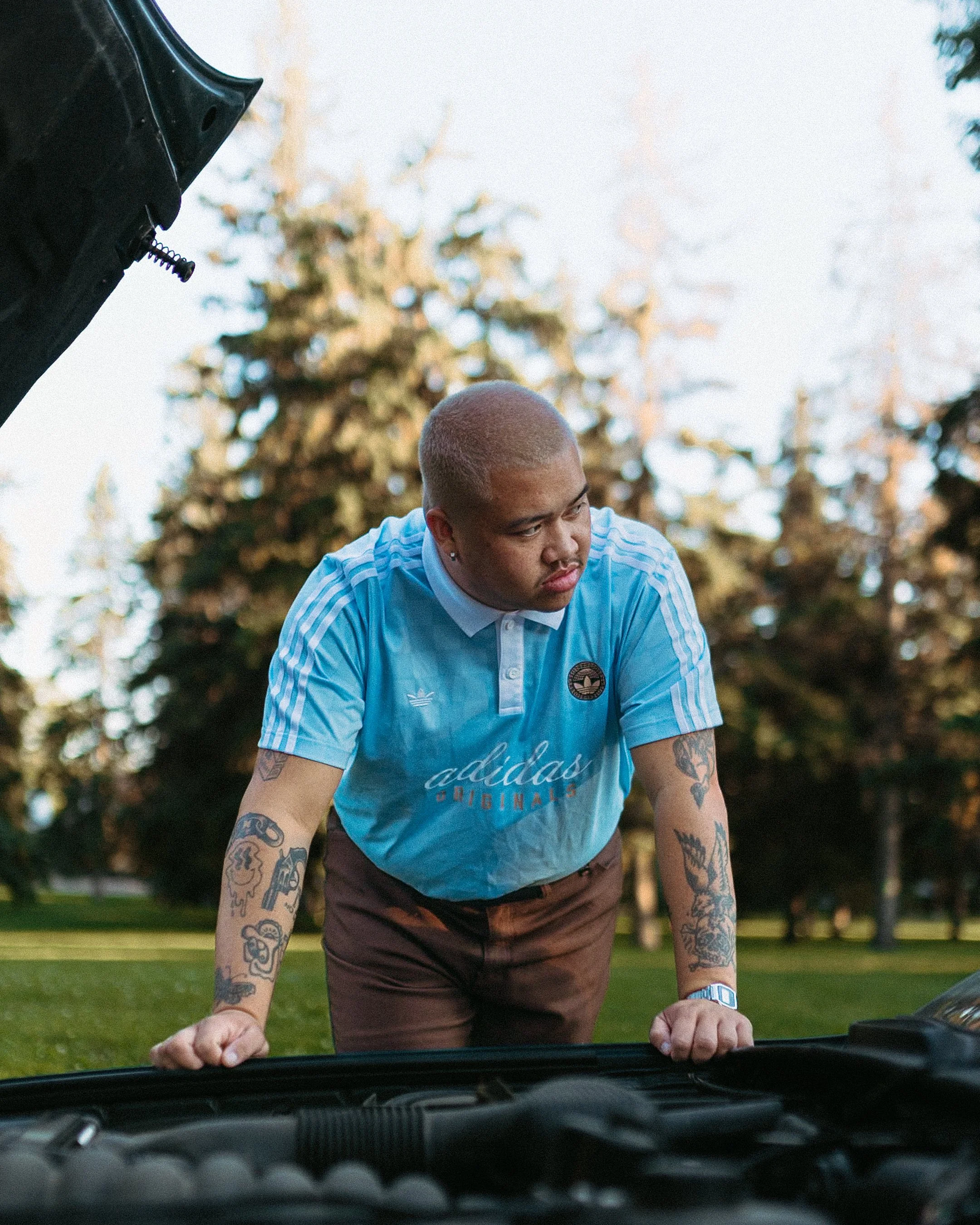 Man with tattoos leaning over the hood of a car, inspecting the engine in a park with trees in the background.