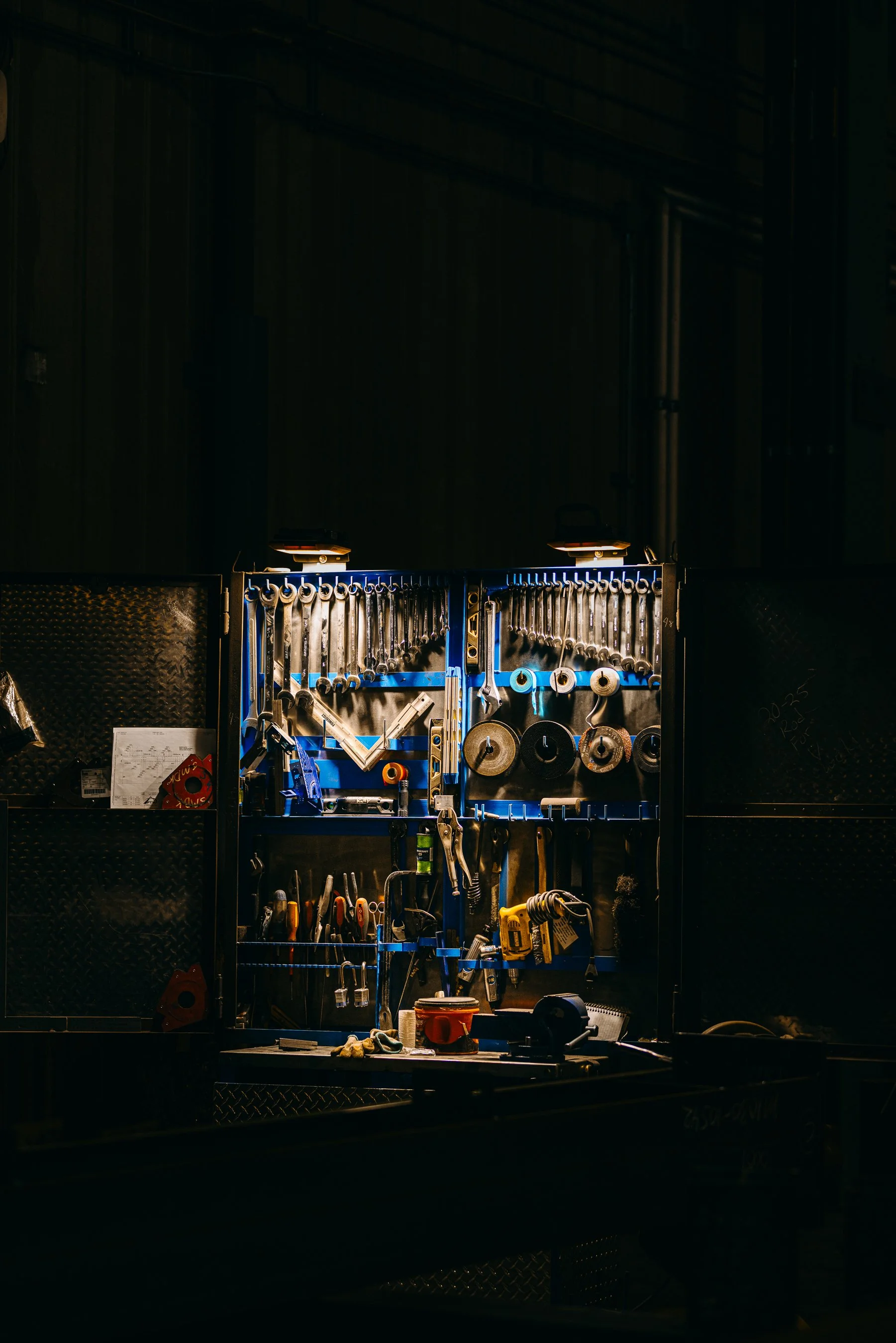 A workbench with tools hanging on a blue pegboard, illuminated by two small overhead lights, in a dimly lit workshop.