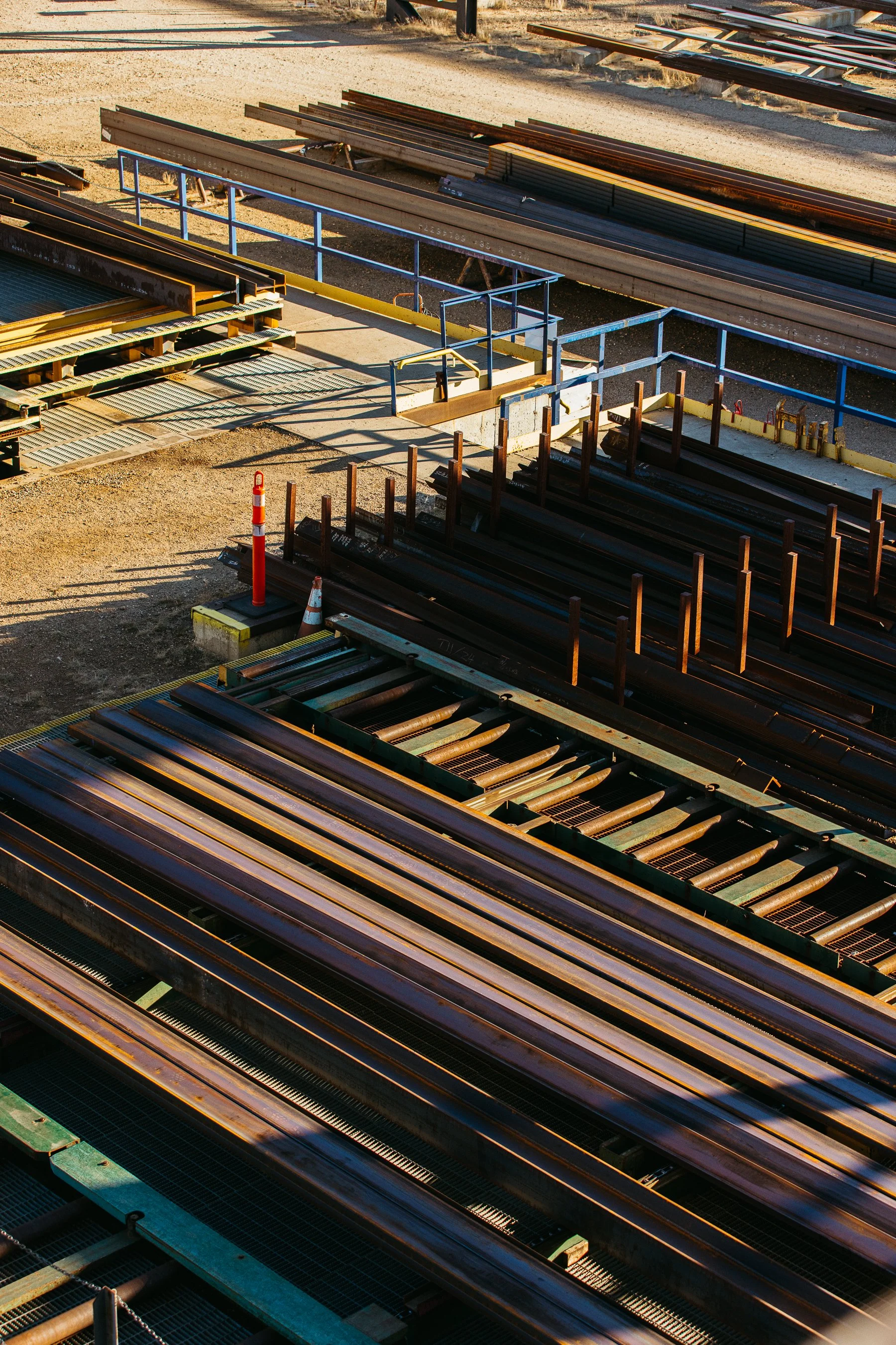 Construction site with steel beams, pipes, and safety barriers, orange traffic cones, and gravel ground.