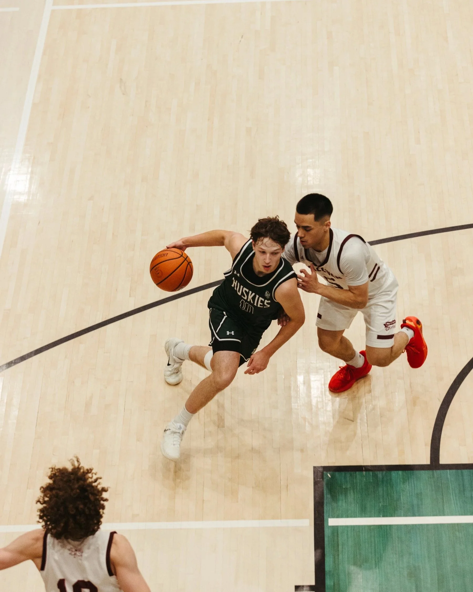 Two high school basketball players in action on the court, one in a black uniform dribbling the ball and the other in a white uniform defending.