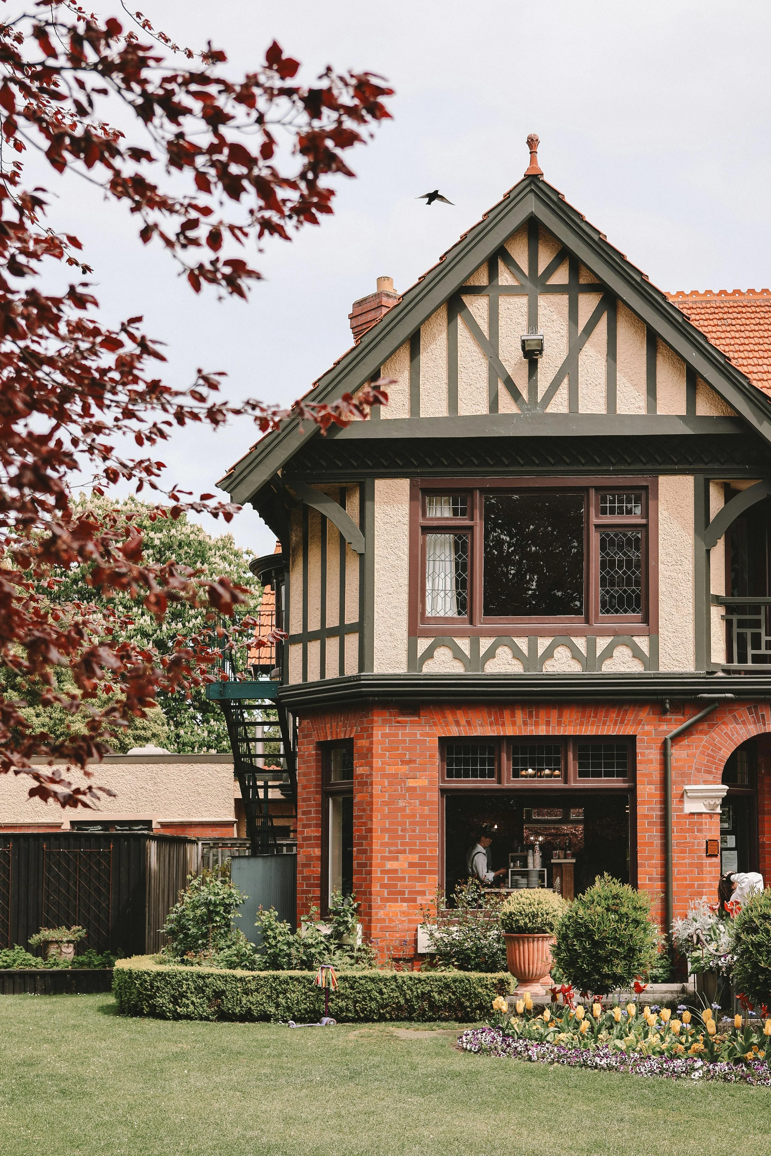 A house with a brick lower level and a Tudor-style upper level with cream and dark wood details. There is a garden in front with flowers, shrubs, and a lawn. A person is inside near the window, and a tree with red leaves is in the foreground. A bird is flying in the sky.