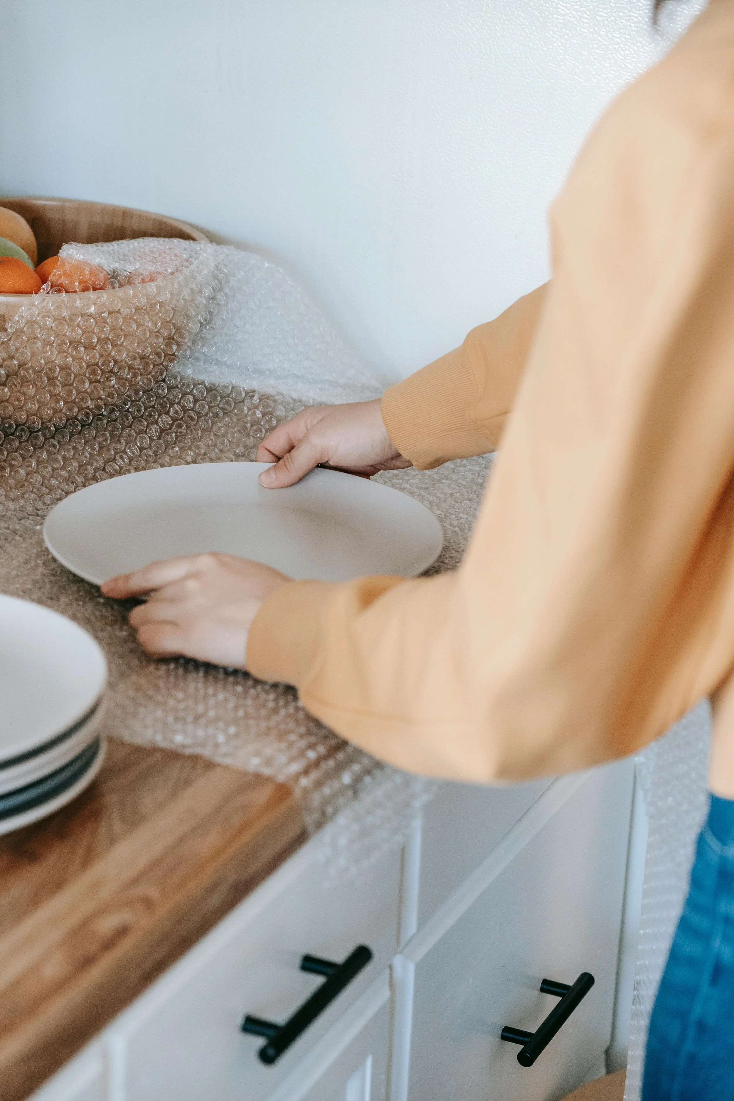 Person setting a white plate on a kitchen counter with a bowl of fruit and stacked plates nearby.