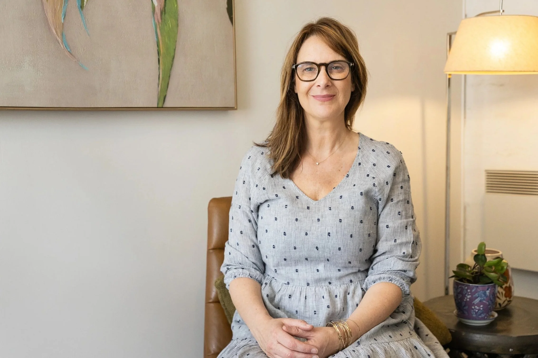 A woman with shoulder-length brown hair, wearing glasses and a gray polka-dot dress, sitting in a room with a painting on the wall and a floor lamp. There are plants in decorative pots nearby.