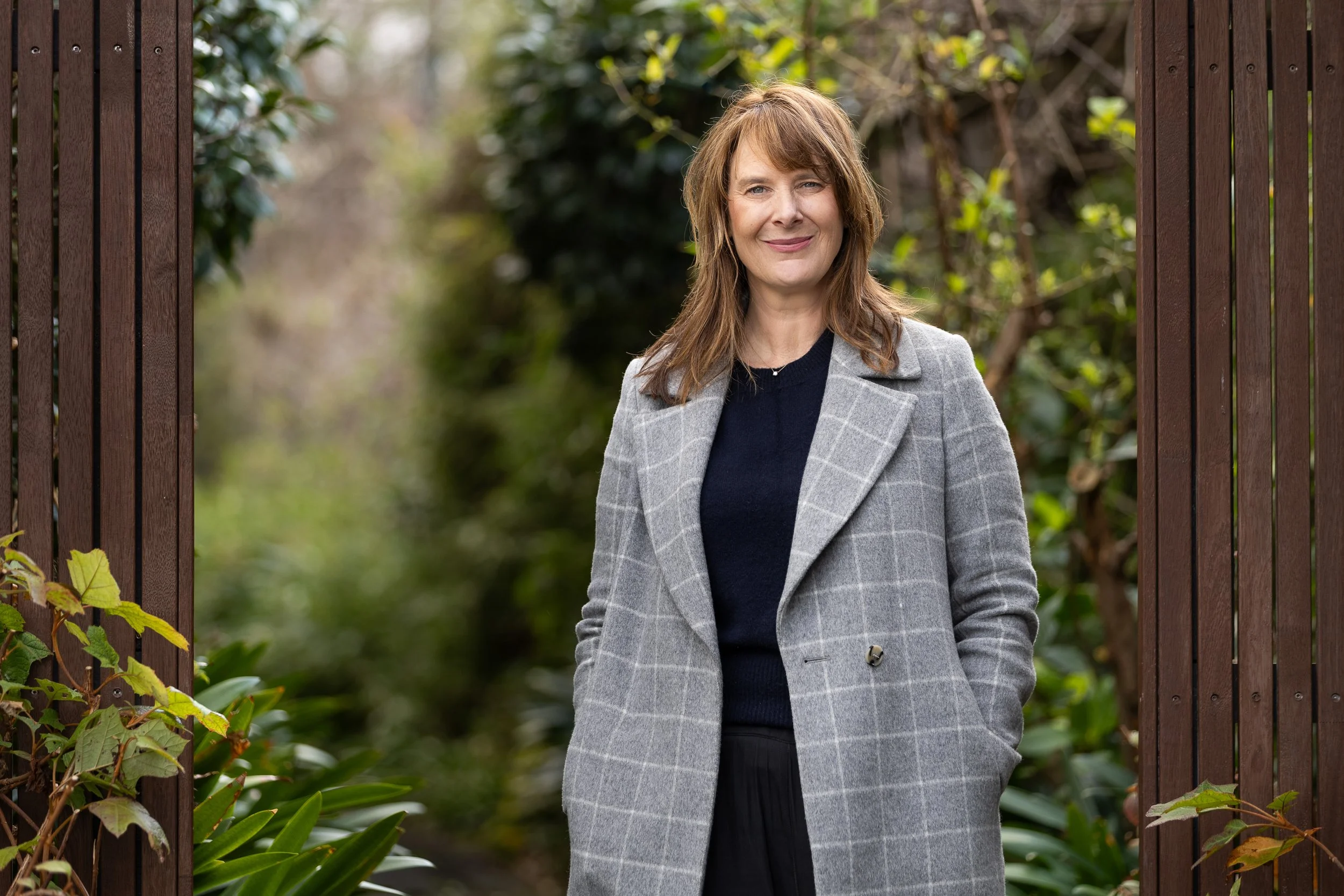 A woman with brown hair wearing a gray checkered coat standing outdoors by a wooden gate with greenery in the background.