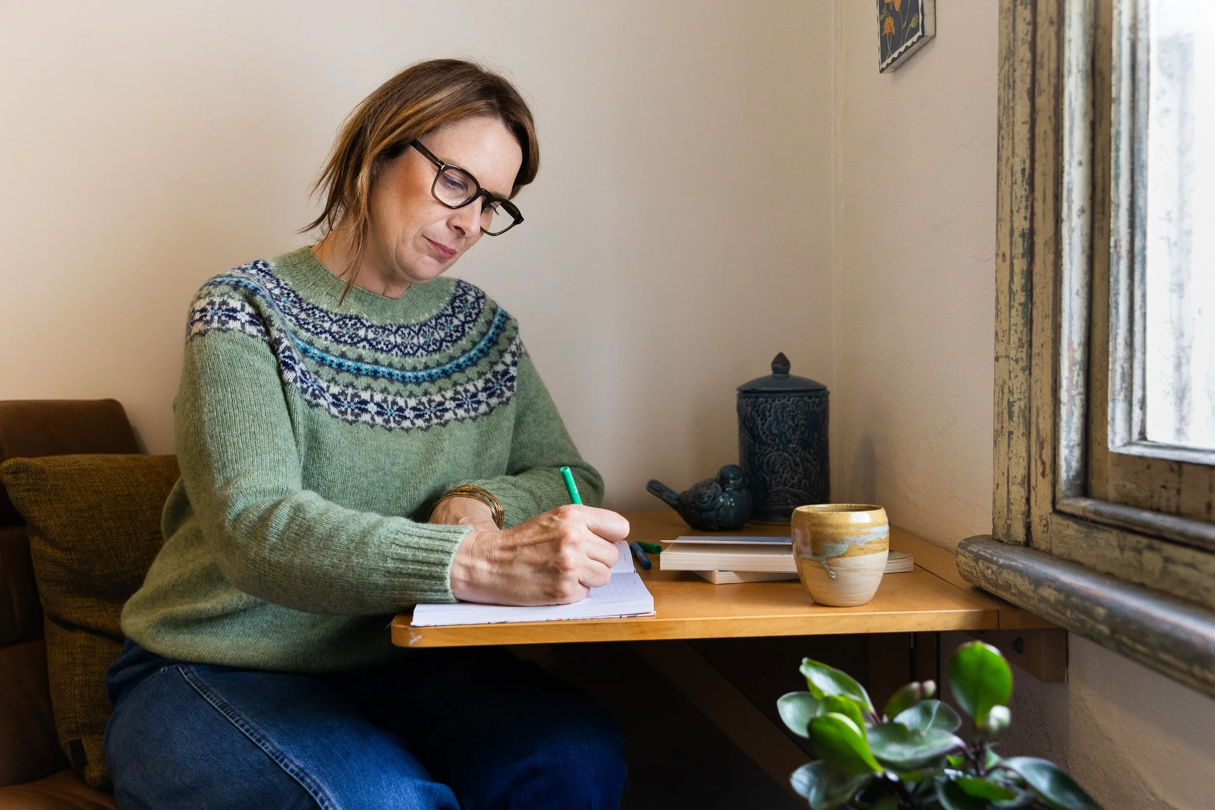 A woman with glasses wearing a green knitted sweater with a blue and white pattern, sitting at a wooden table by a window, writing in a notebook with pens nearby, and decorative pottery on the table.