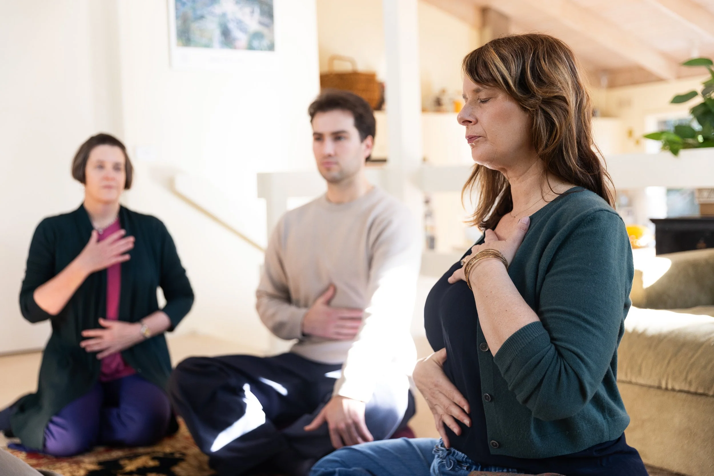 Three people engaged in a moment of prayer or reflection in a cozy, well-lit room, with one woman placing her hand over her heart and the others with hands on their stomachs or chest.