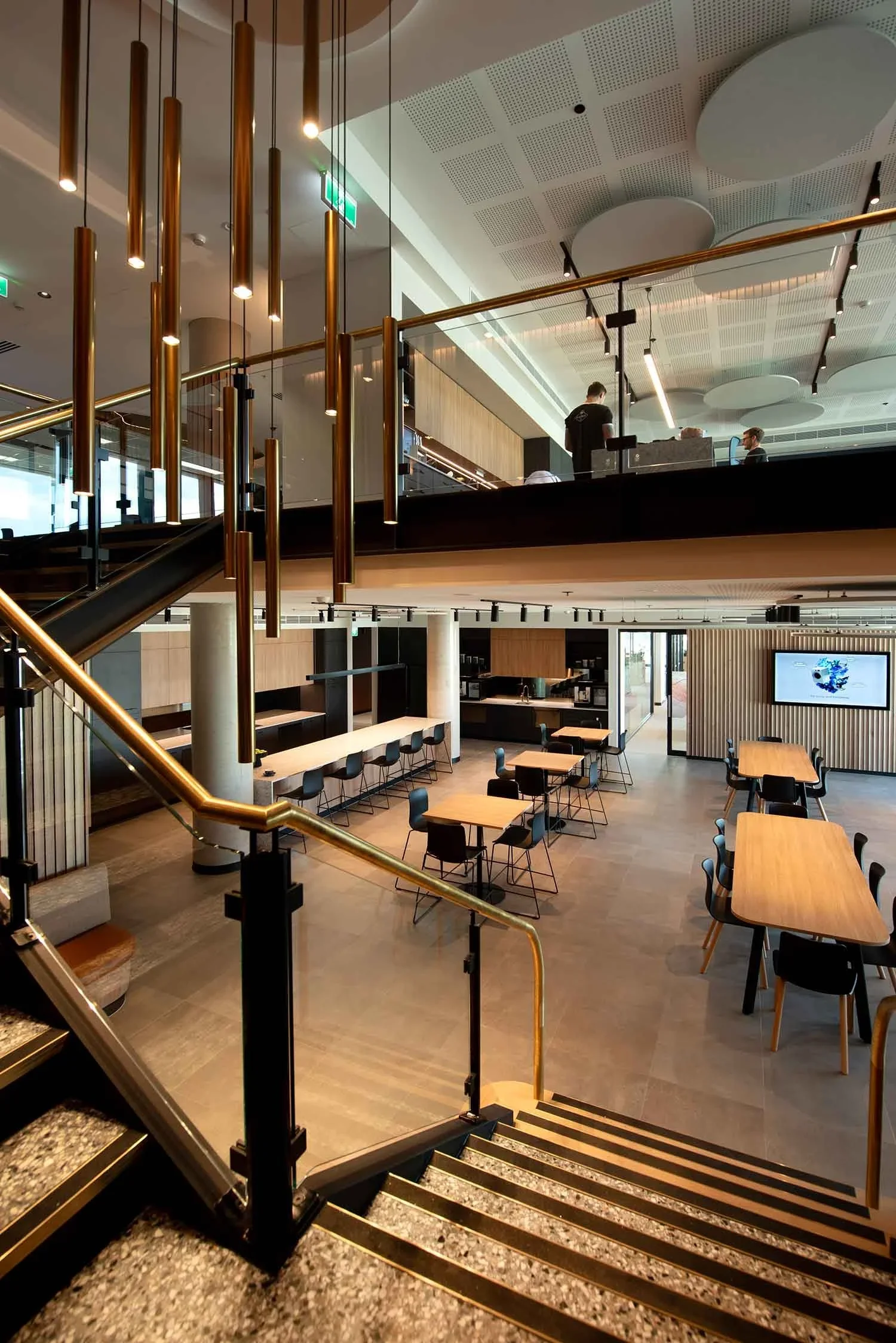 Modern and spacious two-story cafe with wooden tables and black chairs, a staircase with golden bars leading to the upper level, and a flat-screen TV on the wall.