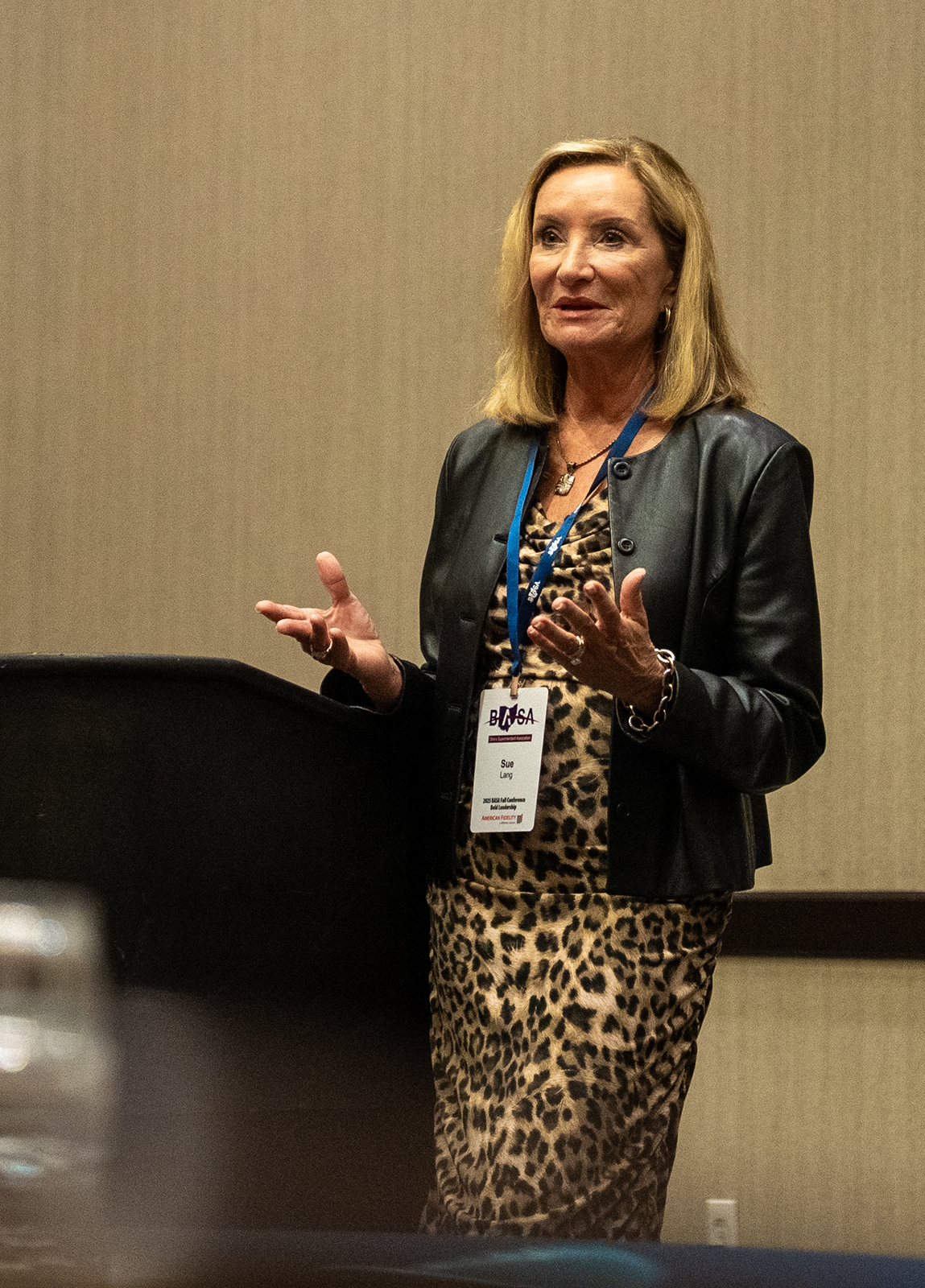 A woman speaking at a conference, wearing a black leather jacket and leopard print dress, with a conference badge around her neck that says Sue Lang.
