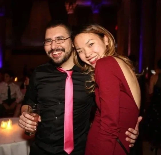 A smiling man and woman are posing together at a social event or party. The man is holding a drink, wearing a black shirt with a pink tie, and glasses. The woman is wearing a red dress, leaning into the man, and smiling.